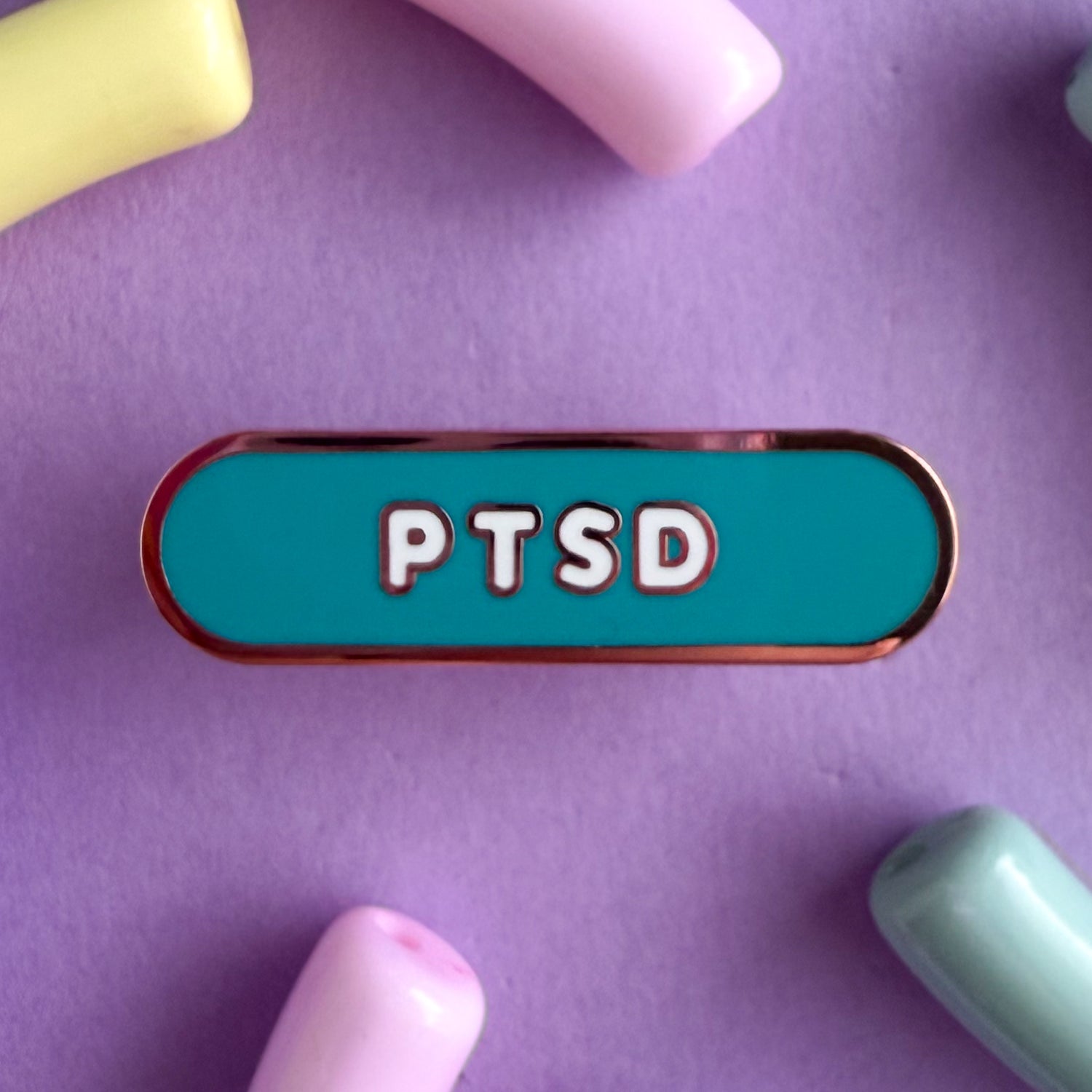 A bandaid shaped pin with the text PTSD in bubble letters. The background is teal and the text is white. The pin is on a purple background.