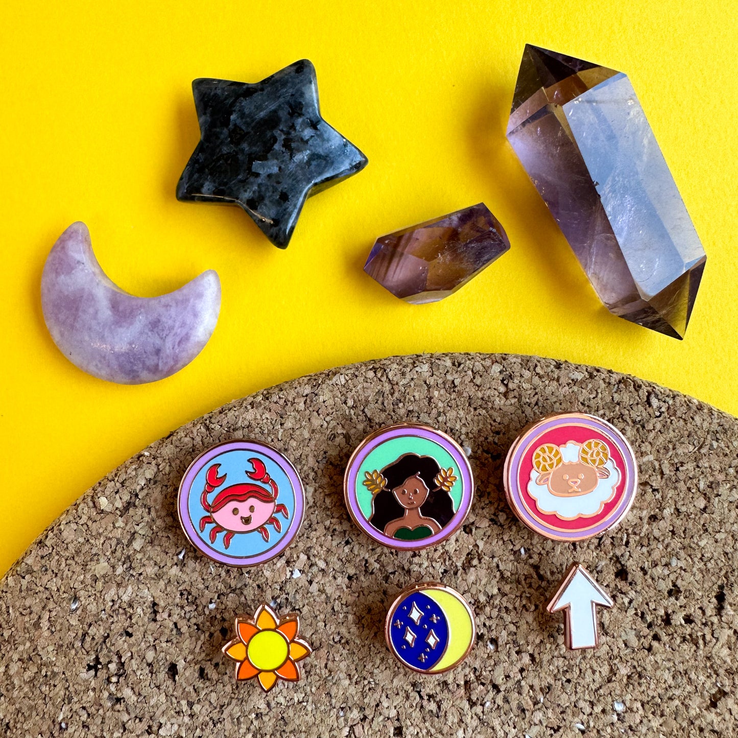 A set of six enamel pins, three are circles with cute illustrations representing zodiac signs. and the other three are shaped like a sun, moon, and rising arrow. The pins are on a corkboard underneath crystals shaped like a moon, sun, and point