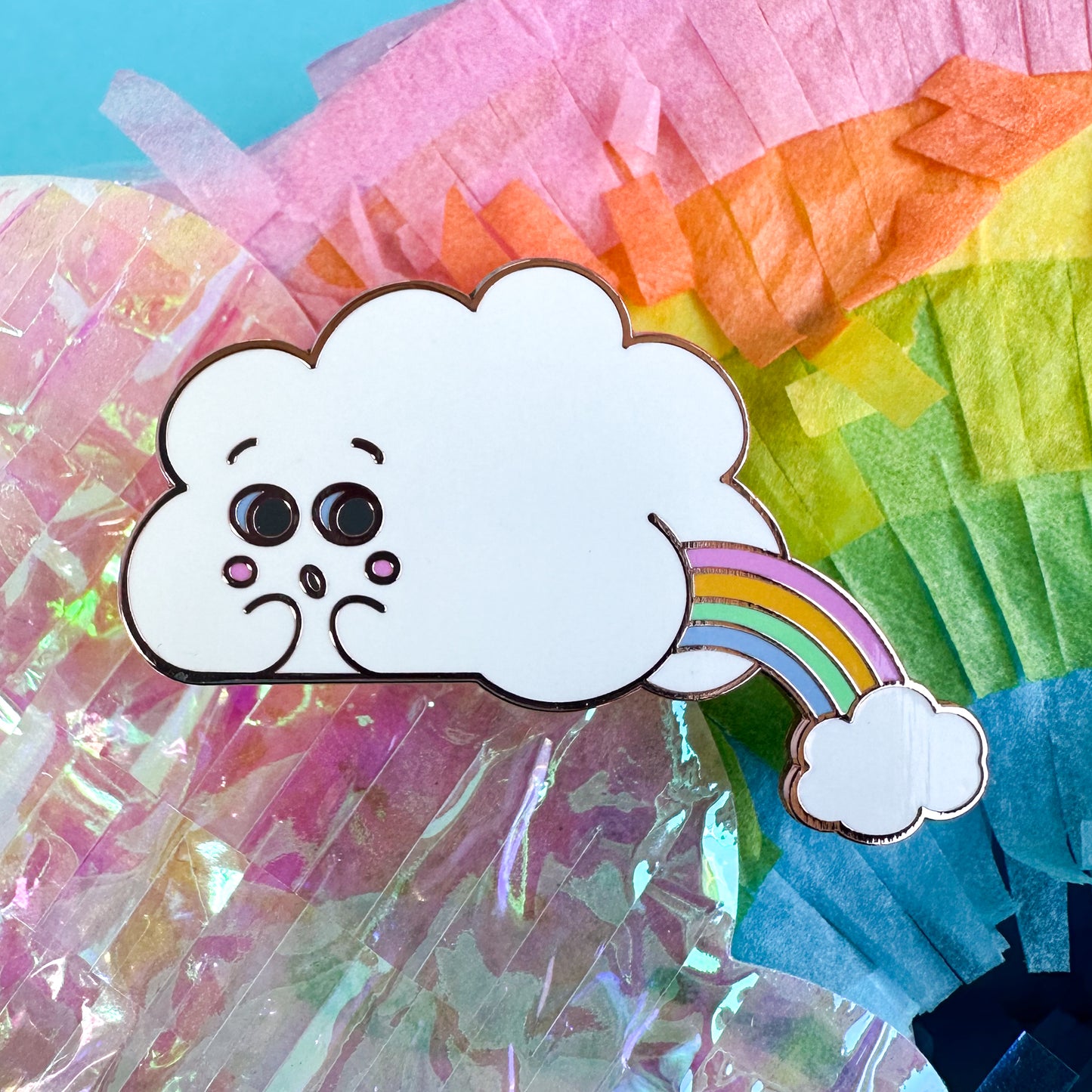 An enamel pin in the shape of a cloud with a face and a butt, there is a pastel rainbow arc coming out of the butt cheeks with another small cloud below it. The pin is sitting on a holographic and rainbow background.