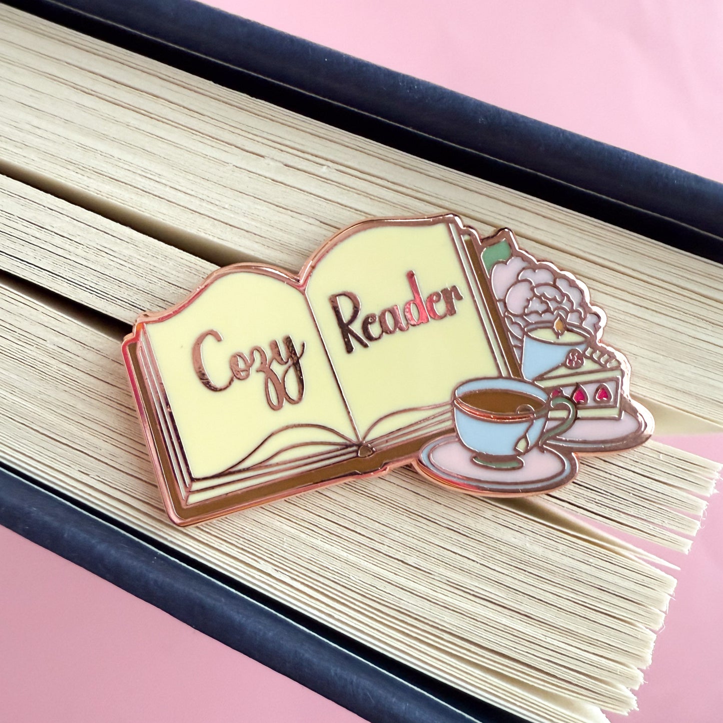 An enamel pin depicting an open book with “Cozy Reader” written across the pages. Beside the book are a full tea cup on a pink saucer, a slice of strawberry cake, a lit candle, and a baby pink peony with a green leaf. The sticker is displayed and the splayed edge of a book.