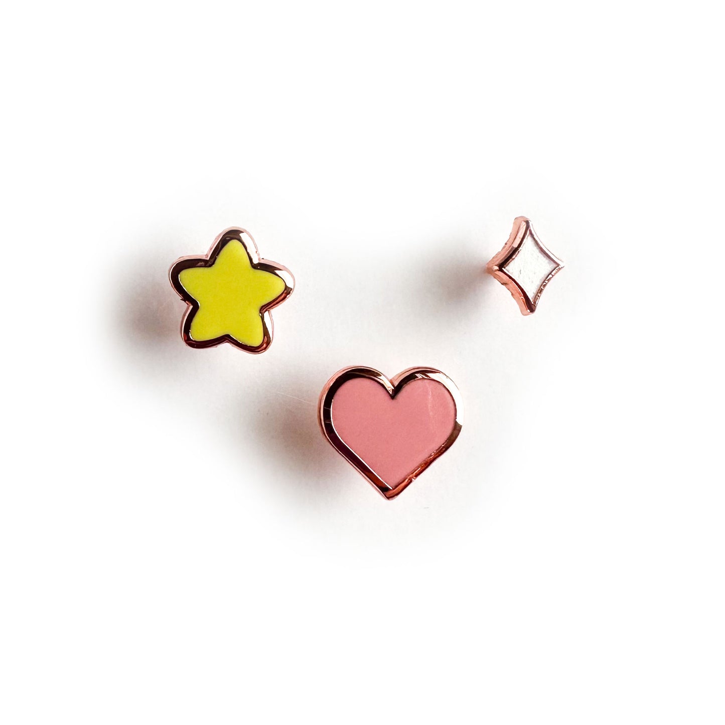 Three small enamel pins one in the shape of a yellow puffy star, one in the shape of a pink heart and one in the shape of a diamond sparkle.
