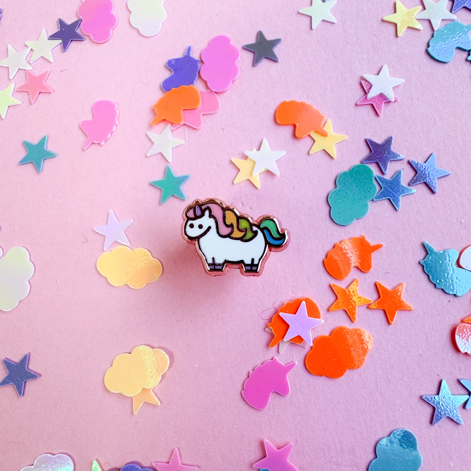 A mini enamel pin shaped like a cute white unicorn with a pastel rainbow mane and tail. The pin is on a pink paper background with pastel confetti around it.