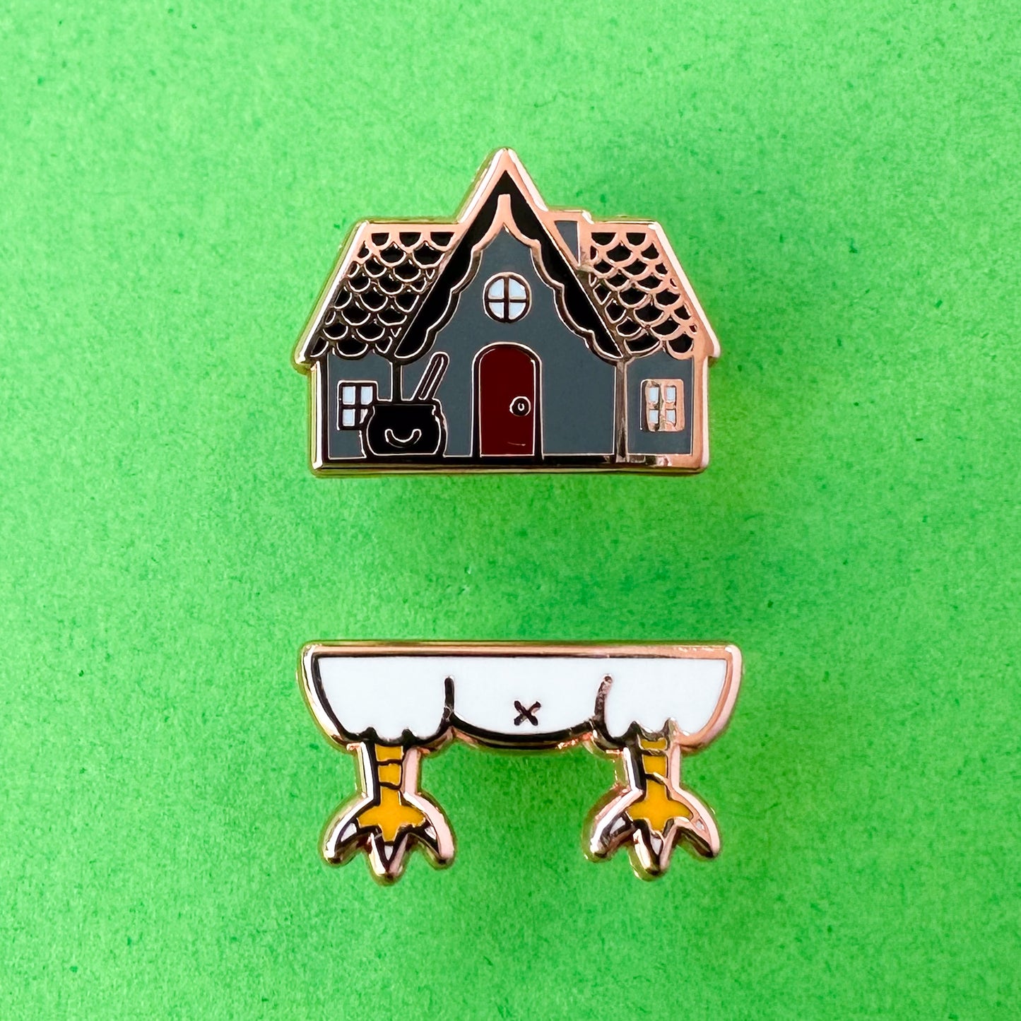 Two pins the top one is a grey house with a gable and a cauldron in front, the bottom pin is chicken legs. The pins are on a green background.