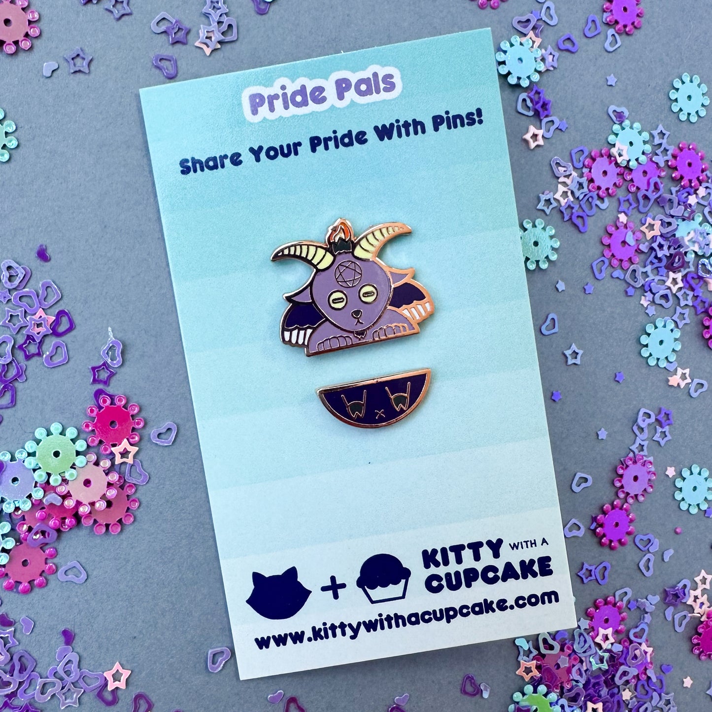 A card that reads "Pin Pals" with two pins representing Baphomet as if he was cut in half horizontally, one pin is the head and torso and the other is the butt and legs. The background is grey with purple, pink and blue confetti glitter around it.