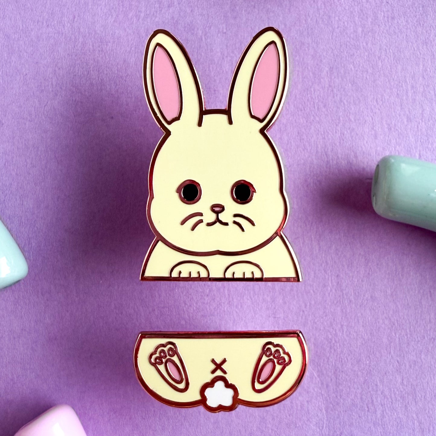 A two piece pin set that comes together to form a white bunny. The top pin is the bunny’s top paws and head with big pink ears. The bottom pin is the bunny’s butt, tail, and feet. The pins are on a purple background.