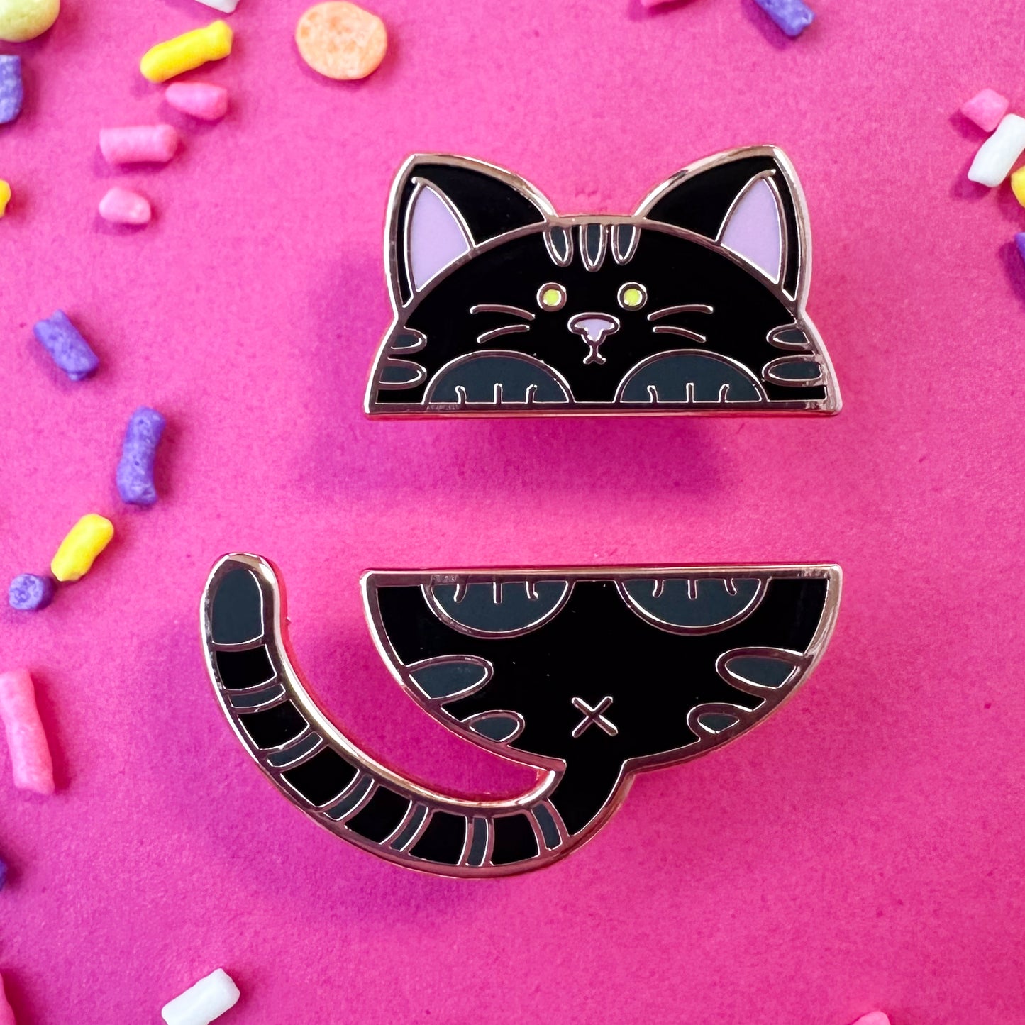 Two enamel pins that come together to form the top and bottom half a of a cute black cat. The pins are on a hot pink background with sprinkles around it.