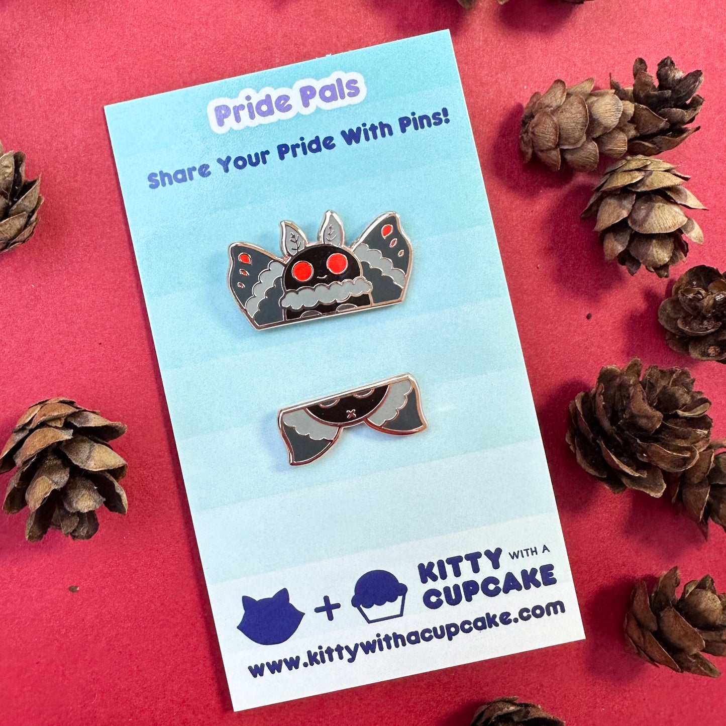Two enamel pins that come together to form a cute illustration of Mothman packaged on a card. The card is on a red background with mini pinecones around.
