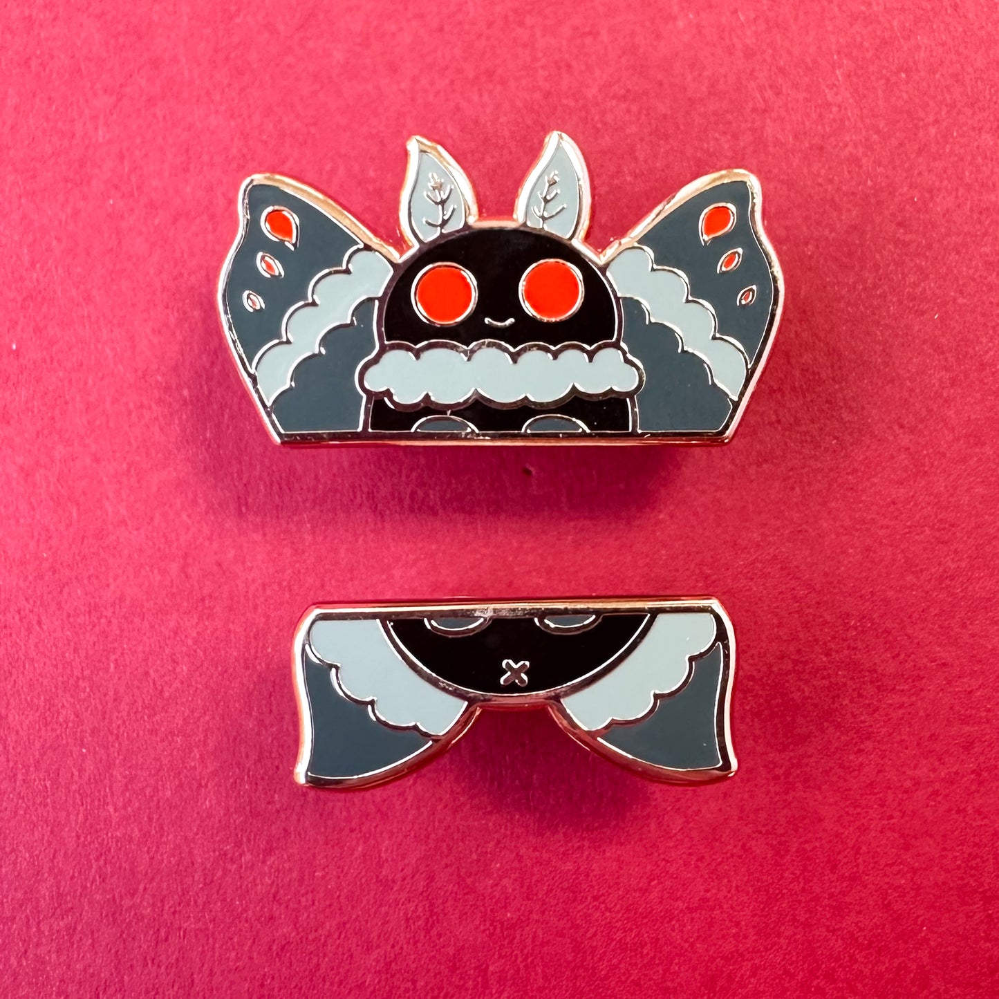 Two enamel pins that come together to form a cute illustration of Mothman. The pins are on a red background.