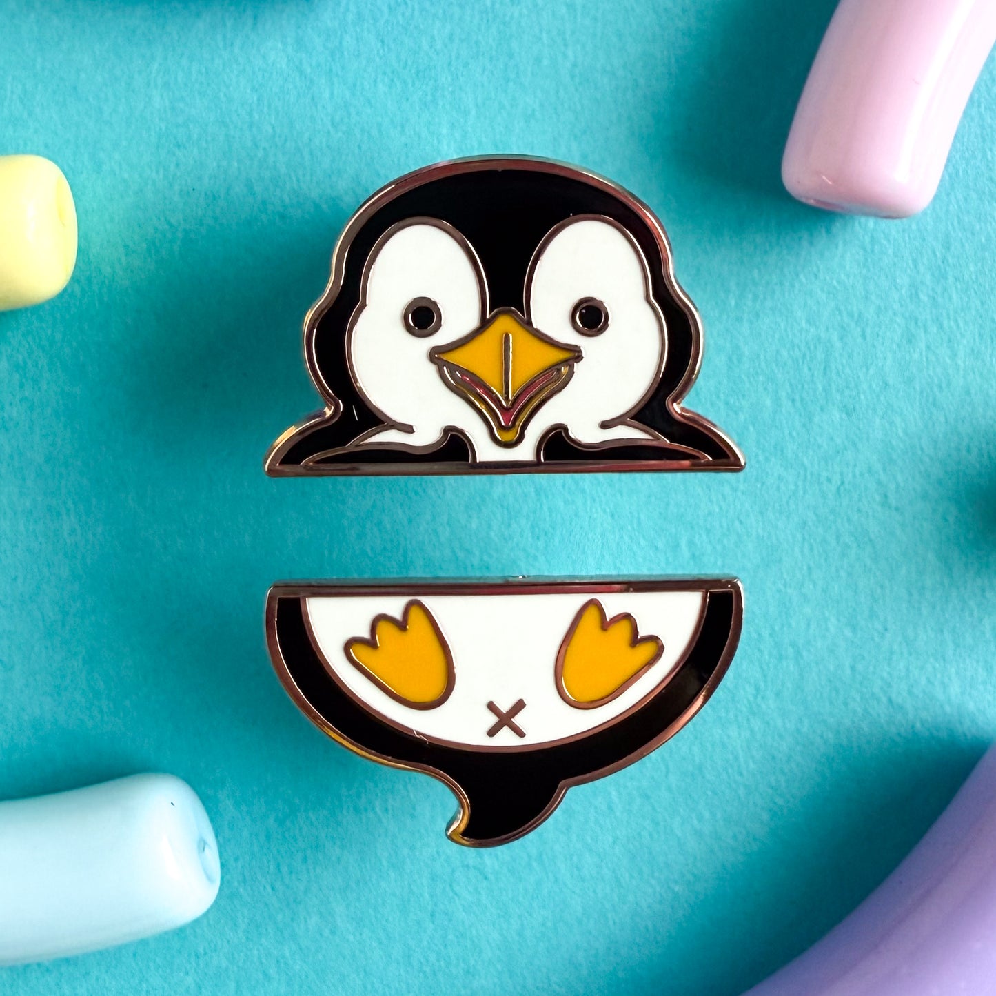 A two piece pin set that comes together to form a penguin. The top pin is the penguin’s wings and head. The bottom pin is the penguin’s butt, tail, and feet. The pins are on a blue background.