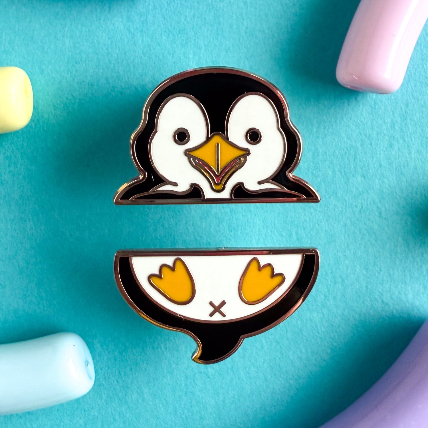 A two piece pin set that comes together to form a penguin. The top pin is the penguin’s wings and head. The bottom pin is the penguin’s butt, tail, and feet. The pins are on a blue background.