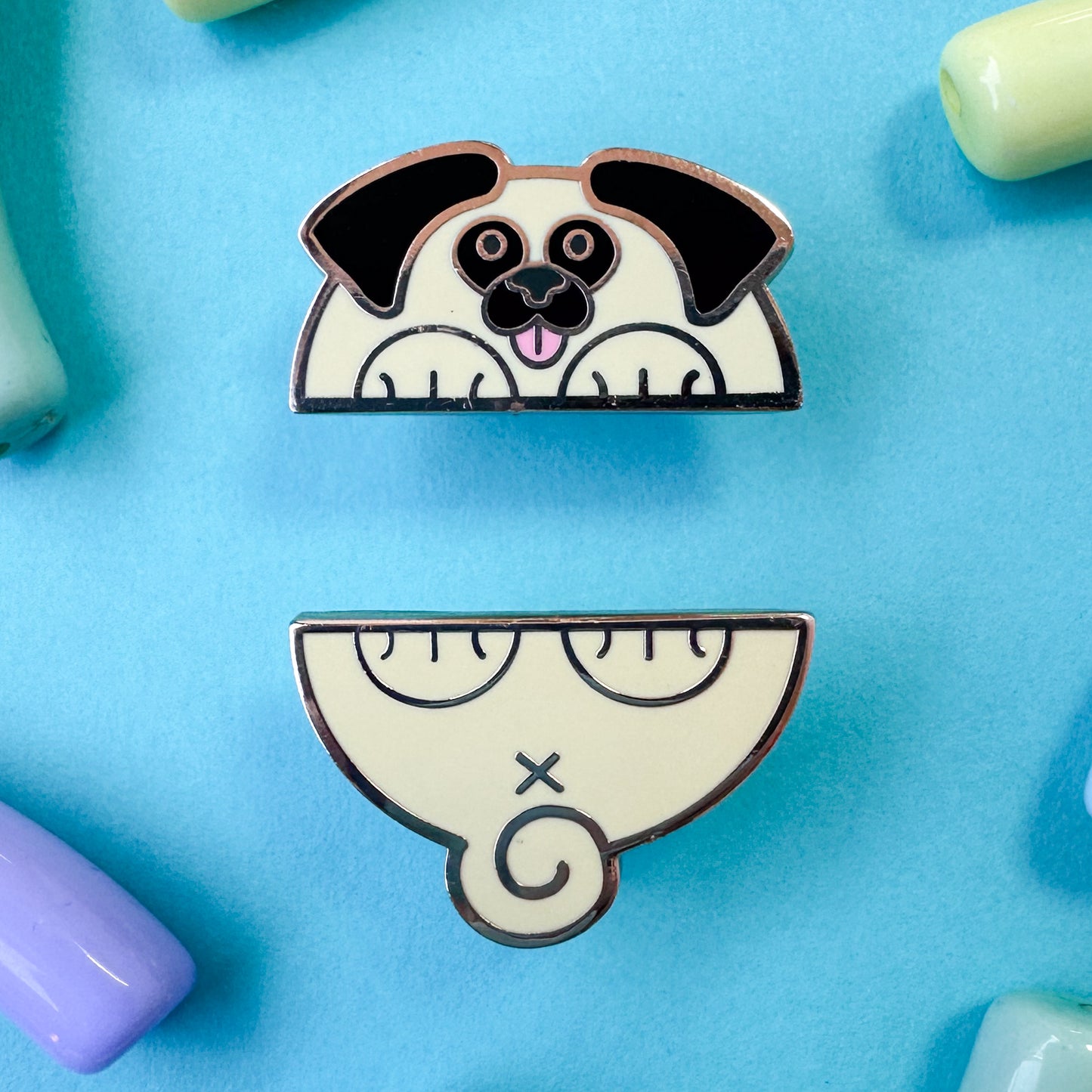 Two enamel pins that come together in the middle to form a pug dog. The pin is on a light blue background with pastel beads around it.