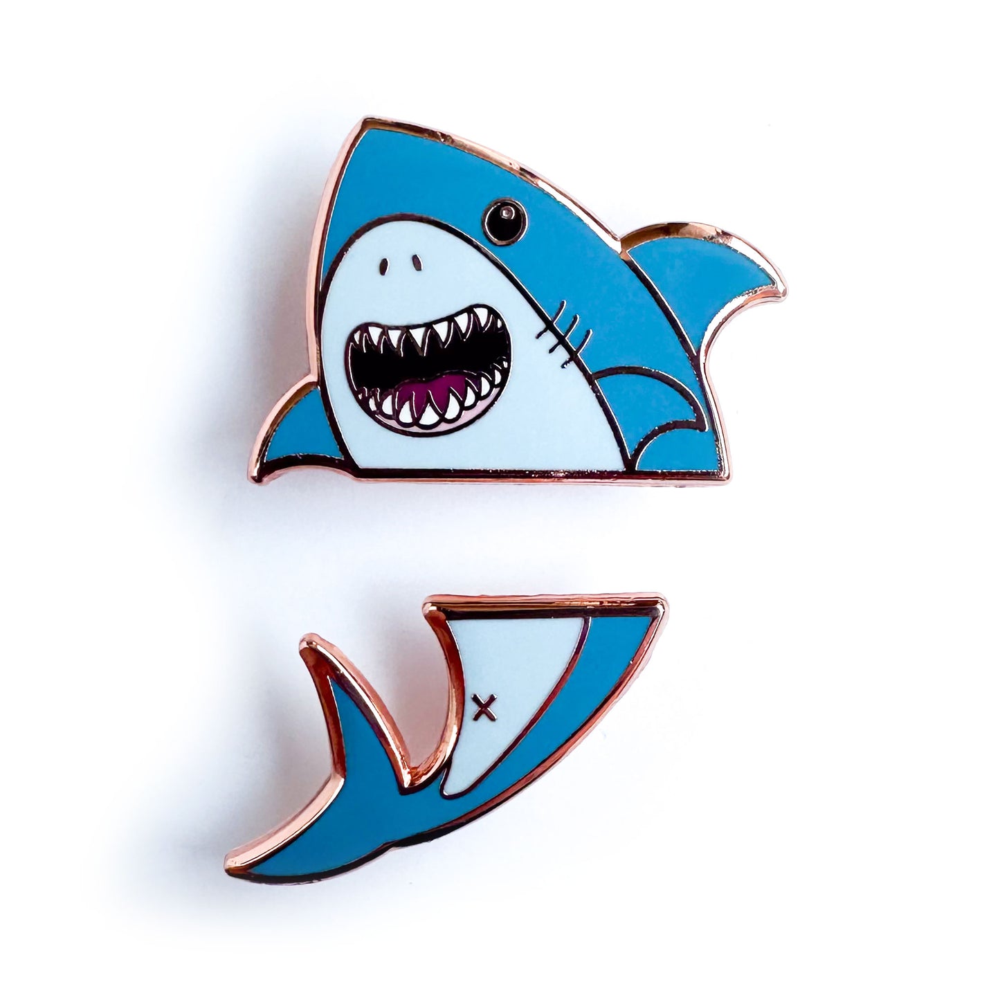 A pair of enamel pins that come together to form a cute illustration of a shark that is split horizontally across the middle. The top pin is the head and side fins, the bottom is the tail.