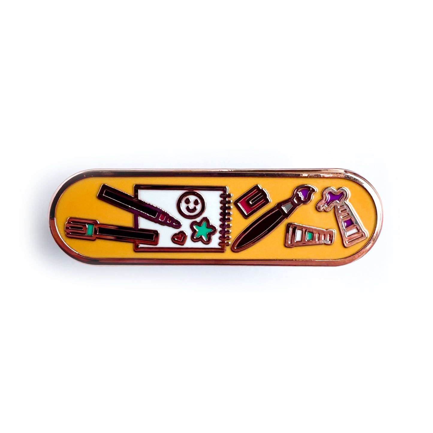 A capsule shaped enamel pin with an orange background and illustrations of art supplies like paints, a notebook, and sketchbook on it.
