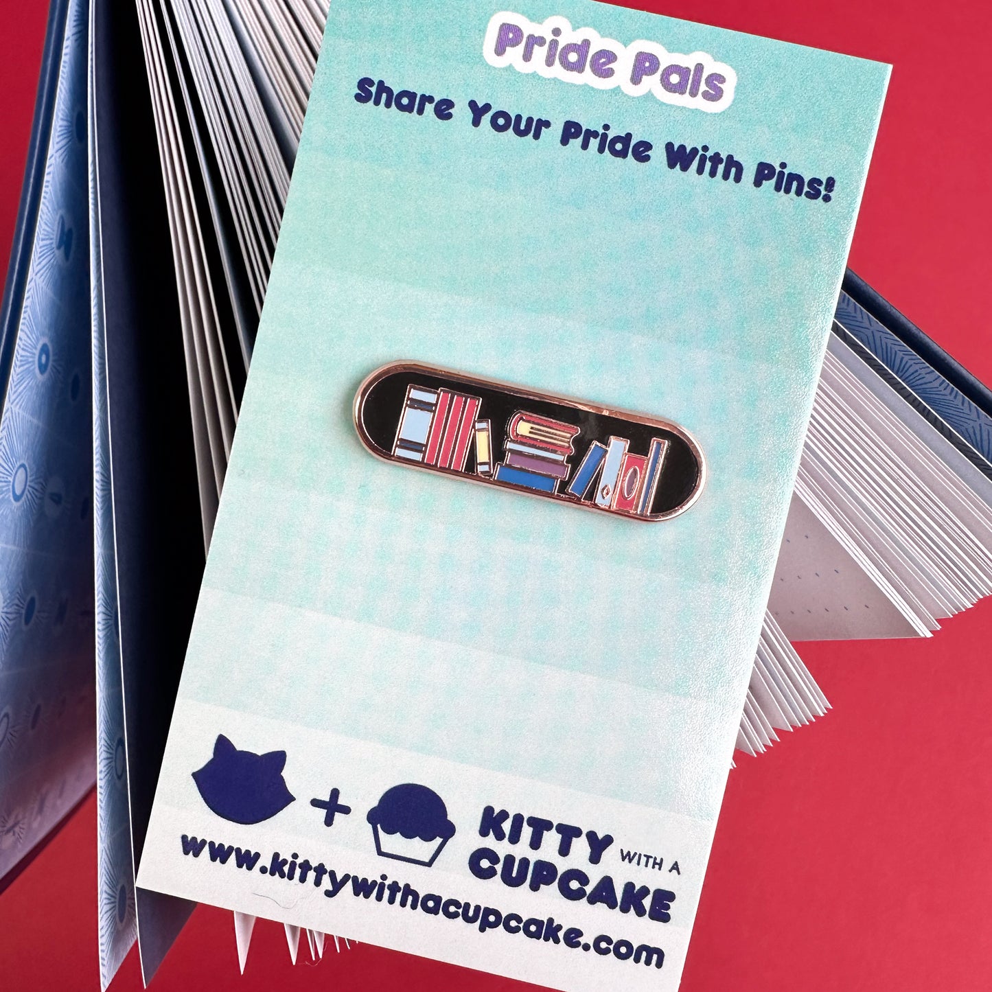 An enamel pin packaged on a card that reads "Pride Pals" the pin has an illustration of a bookshelf on it. 