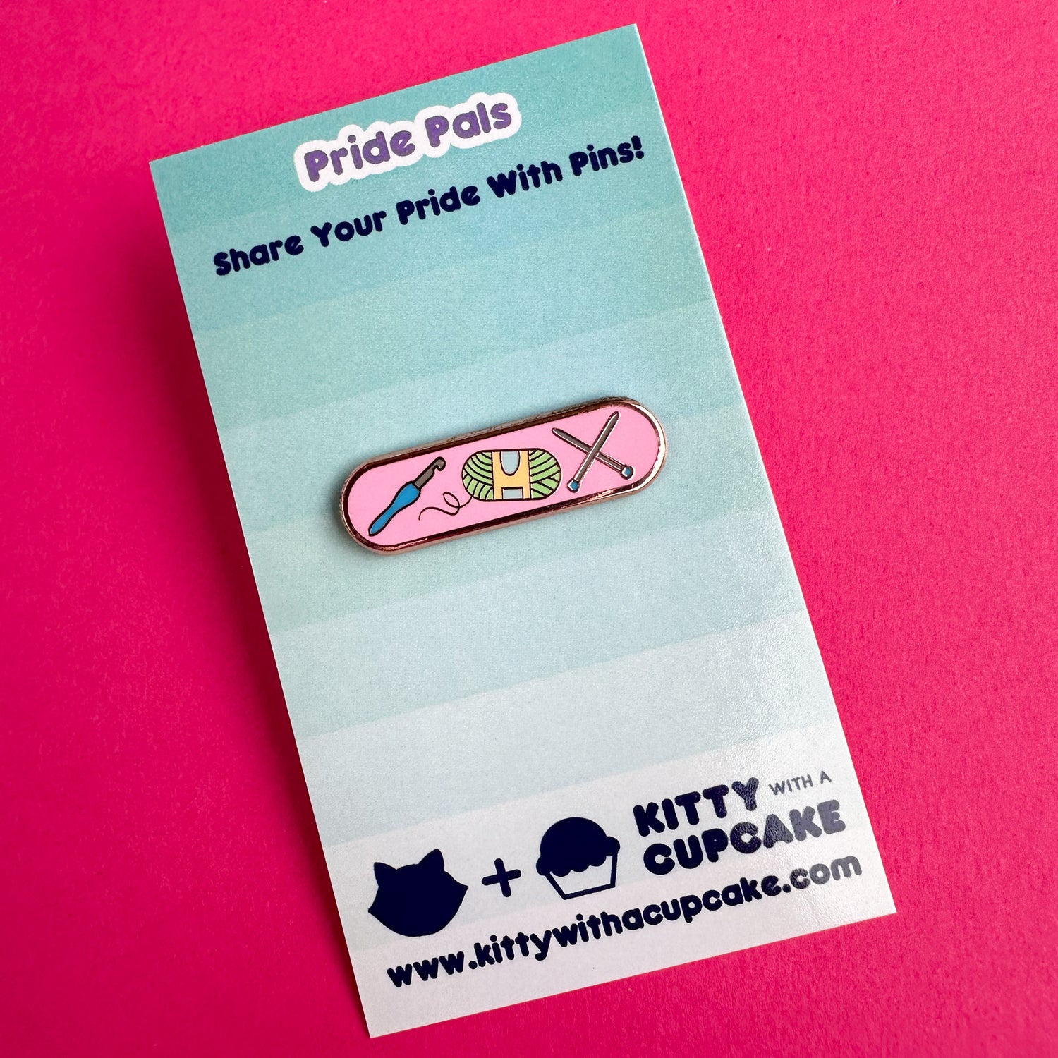 An enamel pin with illustrations of a crochet hook, yarn ball, and knitting needles on it. The pin is packaged on a card and the card is sitting on a hot pink background.