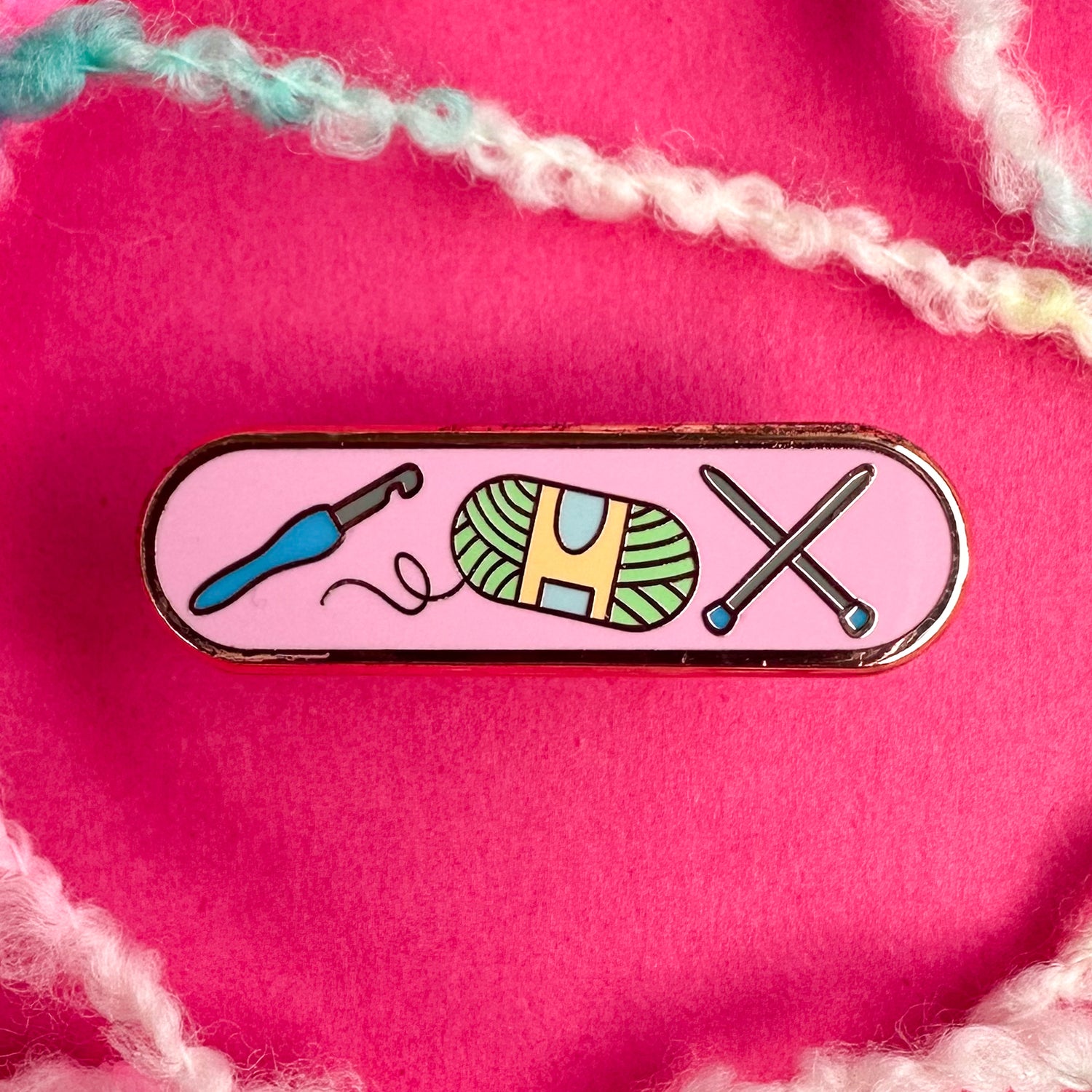 A bandaid shaped pin with a pink background and illustrations of a crochet hook, yarn ball, and knitting needles on it. There is boucle yarn around it and the pin is on a pink background.