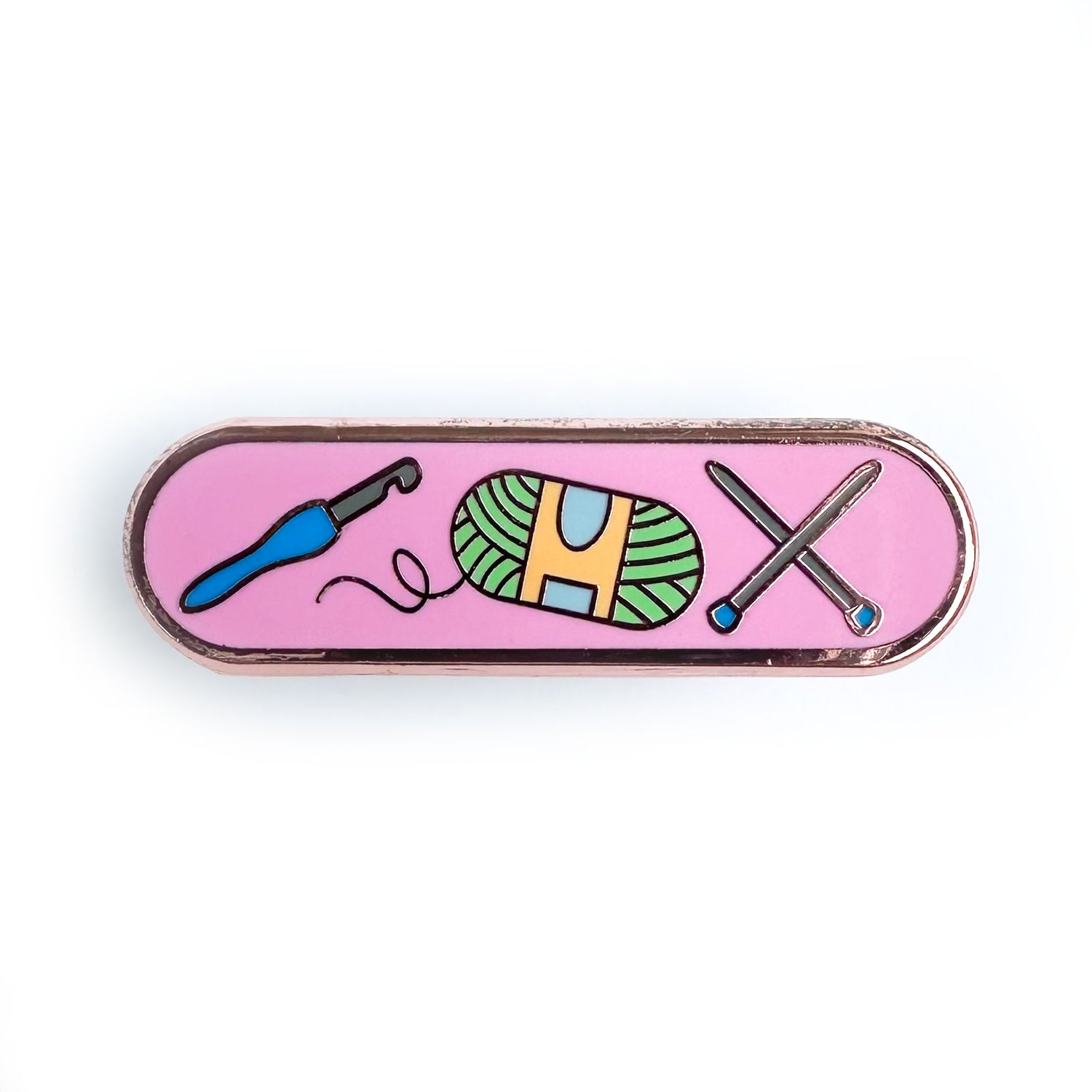 A bandaid shaped enamel pin with a pink background and illustrations of a crochet hook, yarn ball and knitting needles on it.