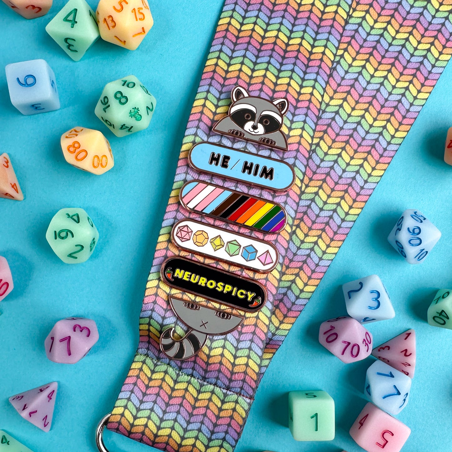 A lanyard with a pastel knit rainbow pattern on it with pins attached to it, there is a raccoon head at the top and his tail at the bottom so it looks like is holding several plaque pins including "He/Him" on a blue background, a pin striped with the colors of the progress pride flag, a pin with pastel rainbow polyhedral dice on it, and a pin that reads "neuronspicy" with peppers on it. There are pastel polyhedral dice around the lanyard on a blue paper background.