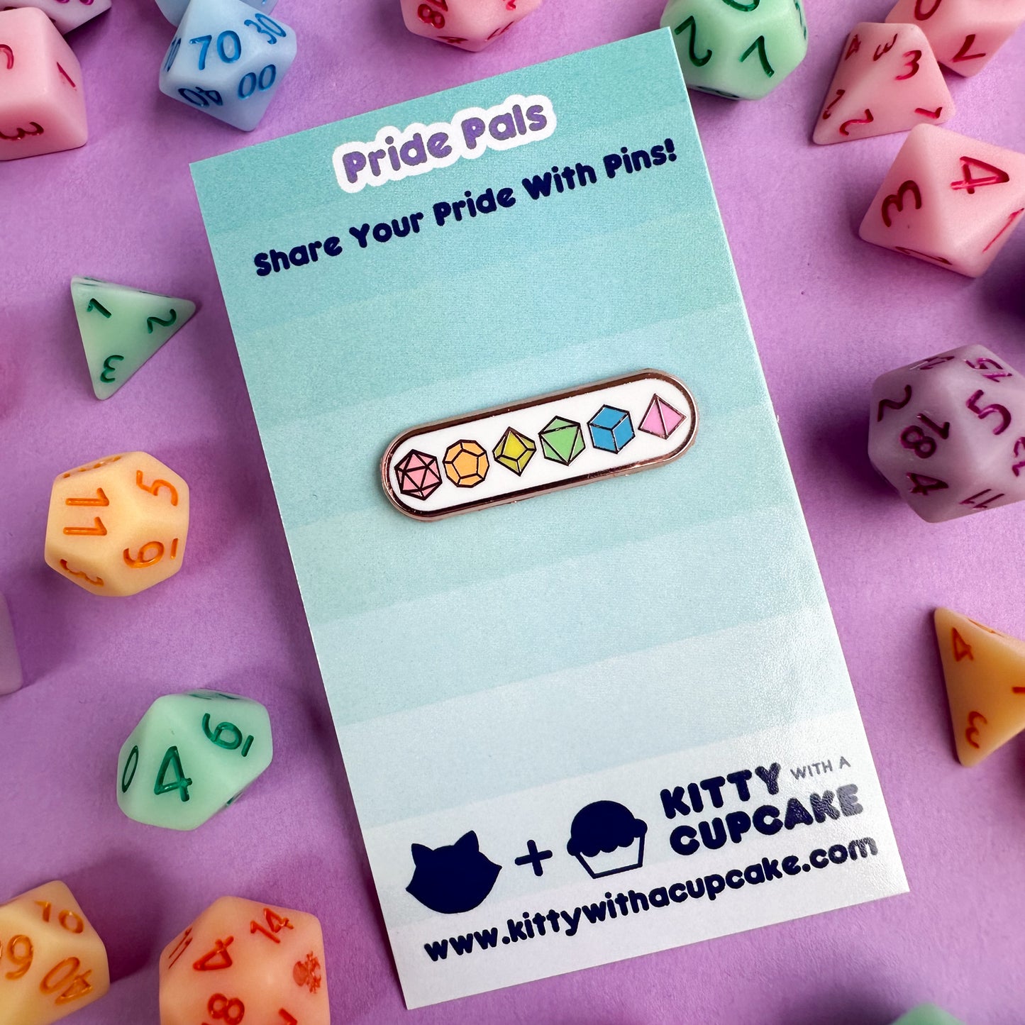 An enamel pin on a card the pin has pastel polyhedral dice on it. There are real pastel polyhedral dice around the card on lavender paper.