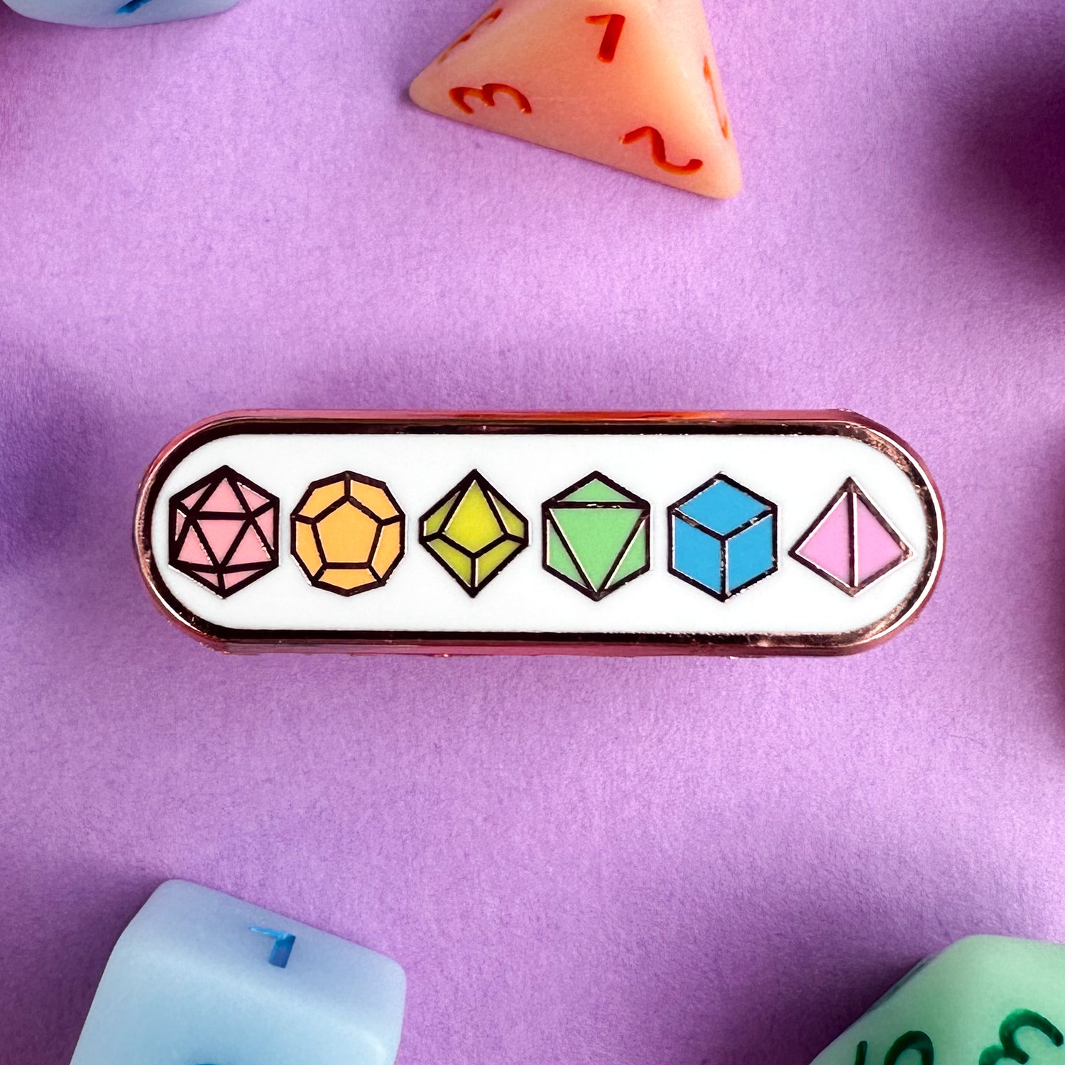 An enamel pin with illustrations of polyhedral dice including a D20, D12, D10, D8, D6 and D4 in pastel rainbow colors. The pin is on a lavender background with real polyhedral dice in the same colors around.
