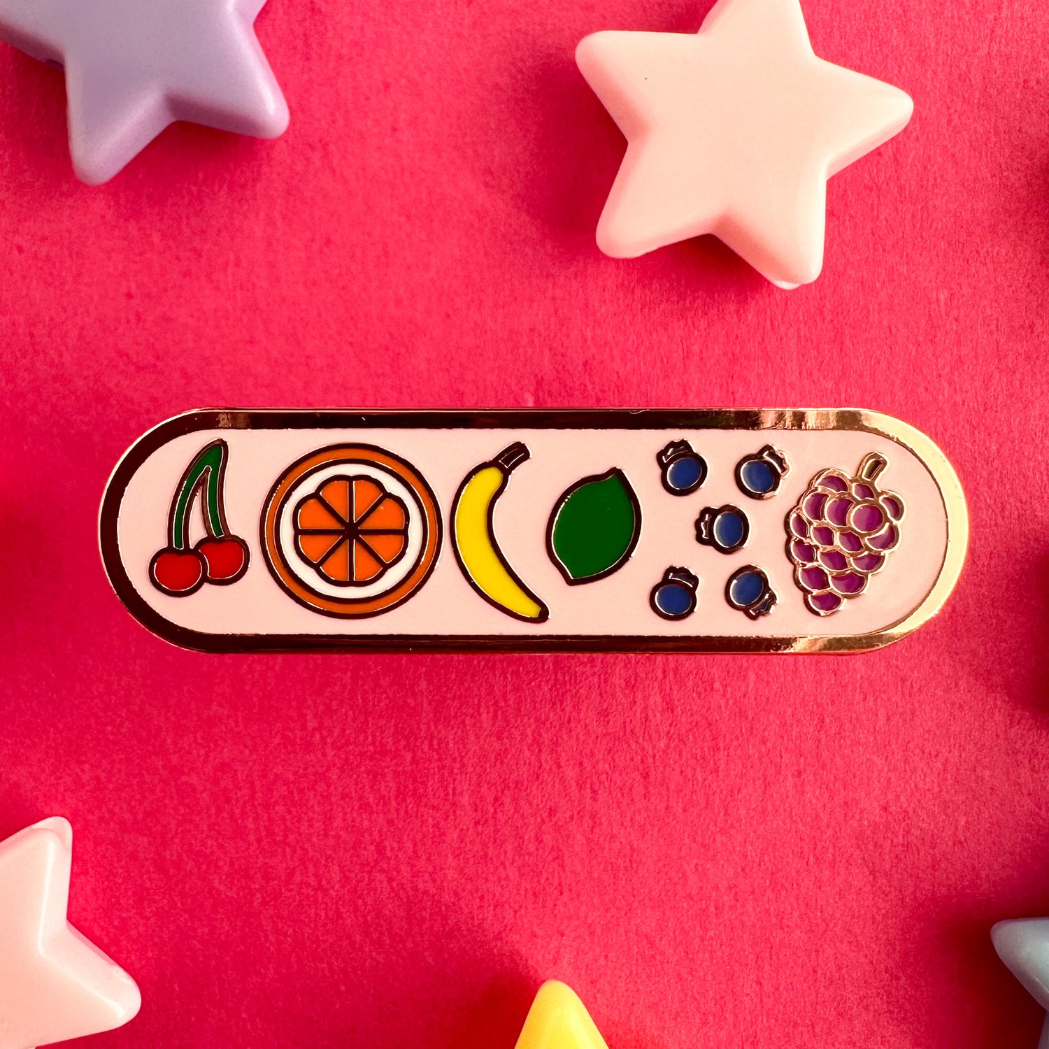 A capsule shaped enamel pin with little doodles of fruit on it arranged in rainbow order. There are cherries, an orange, a banana, a lime, blueberries, and grapes. The background of the pin is light pink. The pin is on a hot pink backdrop with plastic star beads around it.