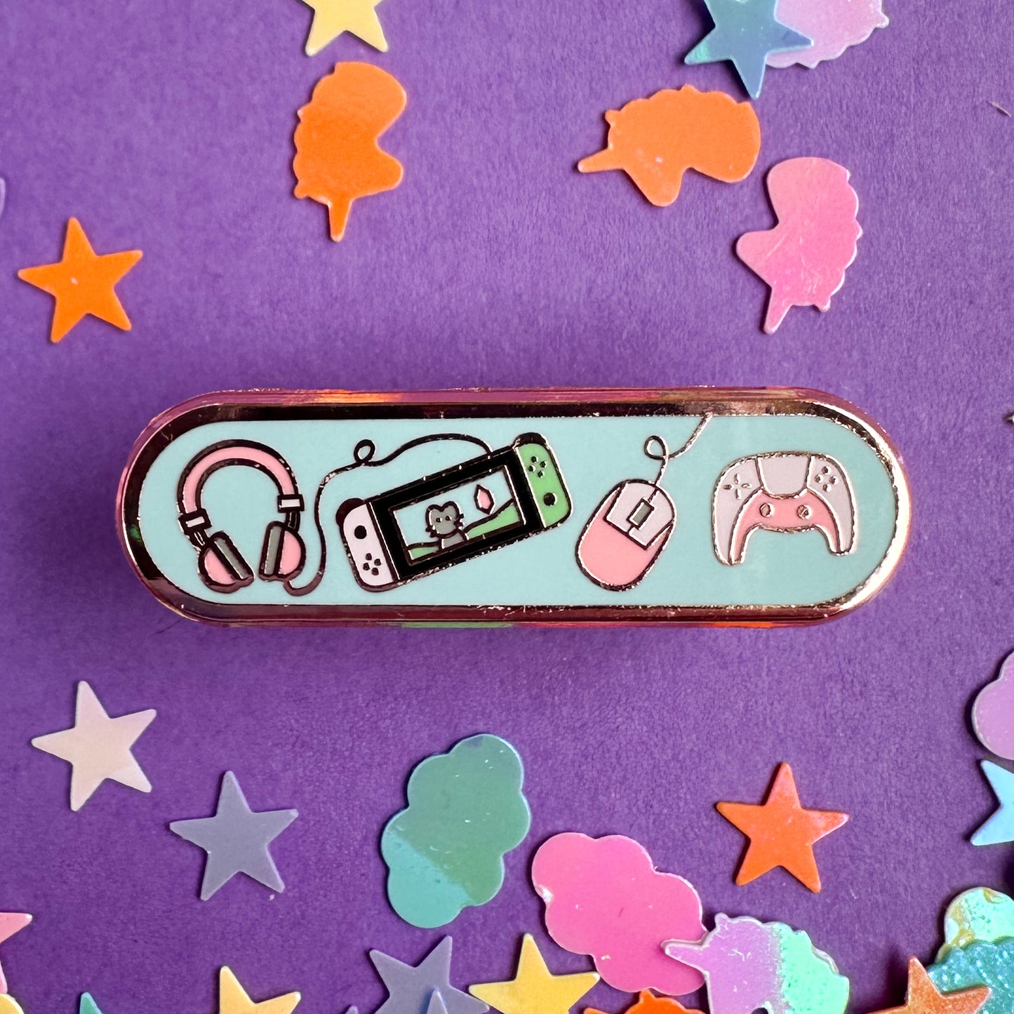 An oval shaped pin with illustrations of video game system equipment on it. The pin is on a purple background with pastel unicorn and star glitter around it.