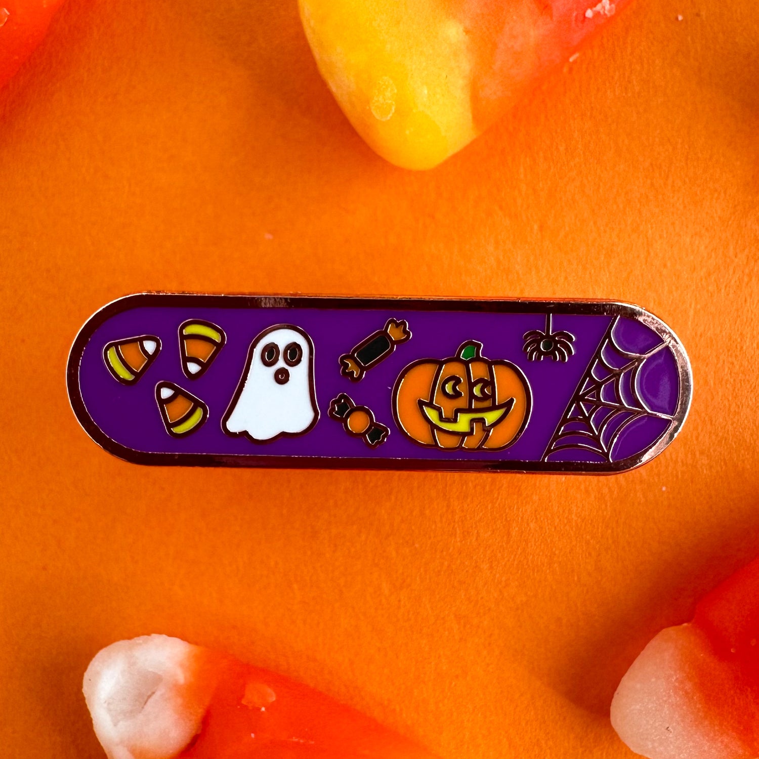 A capsule shaped enamel pin with a purple background and cute illustrations of candy corn, a ghost, candy, a jack o lantern, and a spider on it.  The pin is on an orange paper backdrop surrounded by candy corn.