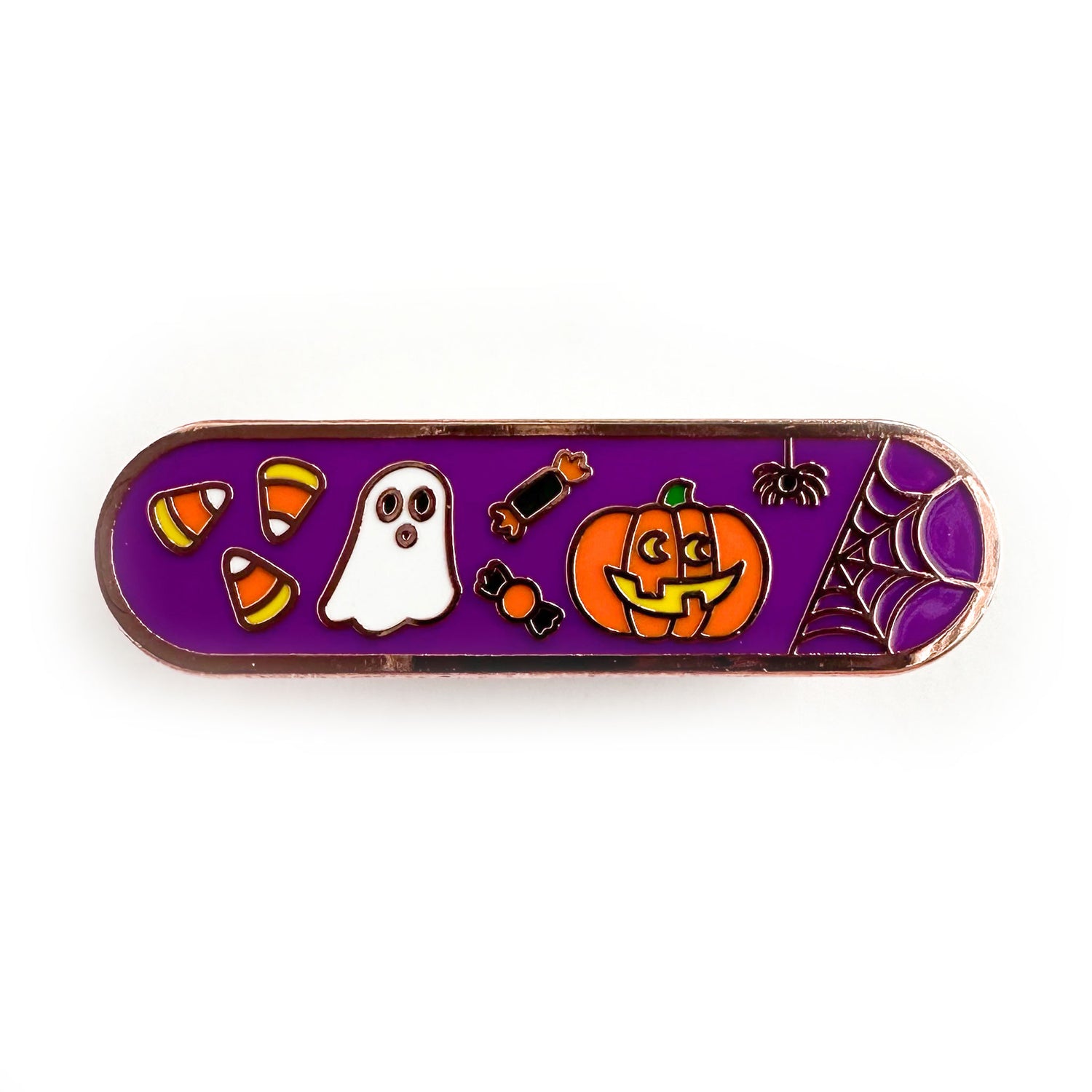A capsule shaped enamel pin with a purple background and cute illustrations of candy corn, a ghost, candy, a jack o lantern, and a spider on it.