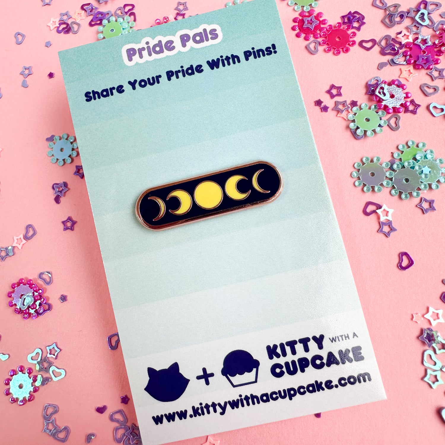 A capsule shaped pin with moon phases on it packaged on a card. The card is on a pink background with blue and pink glitter around it.