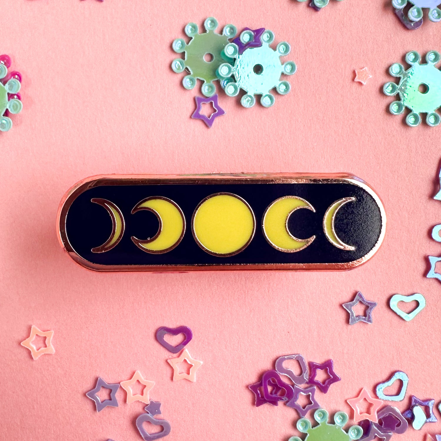 An oval pin with a full moon in the center and crescent moons framing it. The pin is on a pink background with glitter around it.