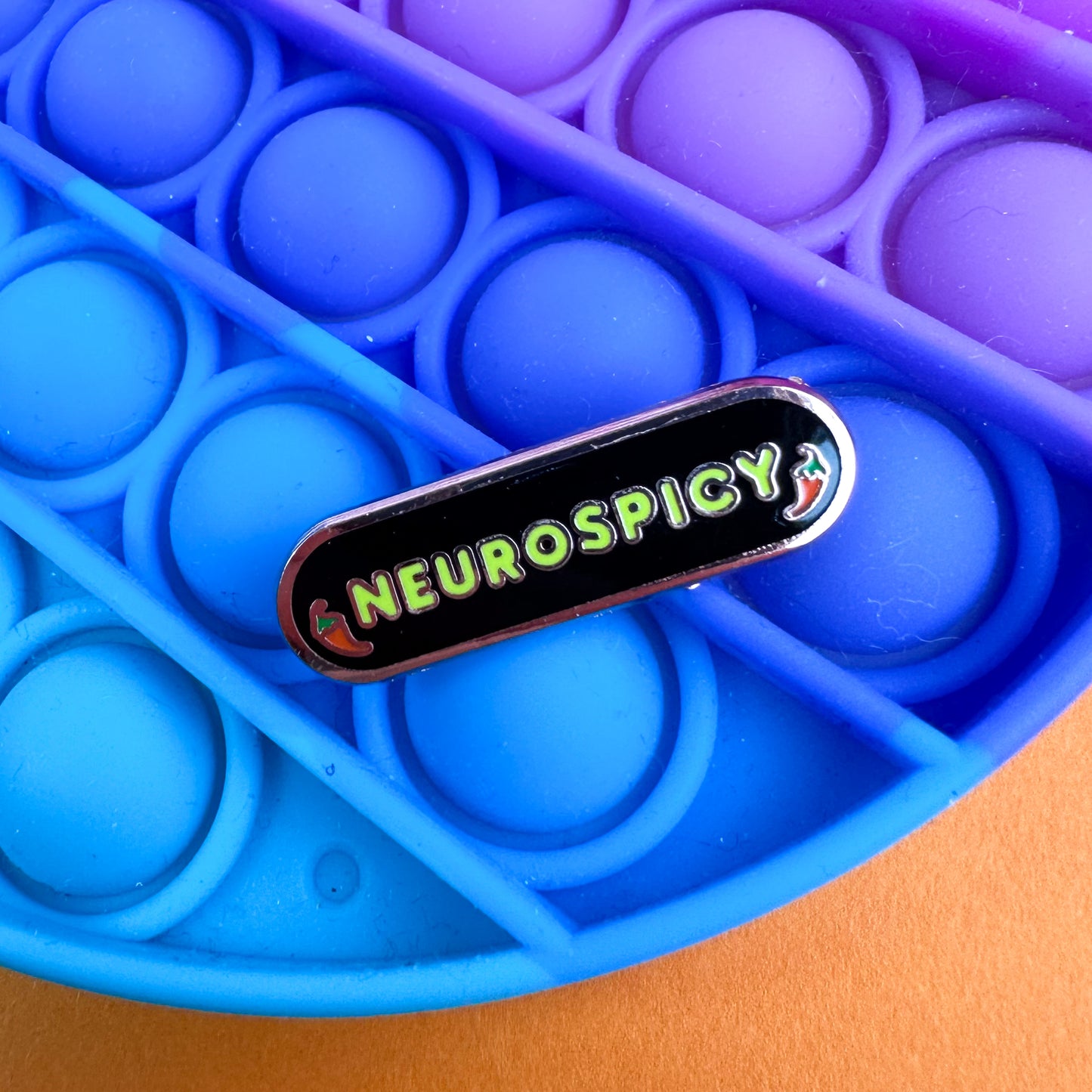 A capsule shaped enamel pin that reads "Neurospicy" with chili peppers around it on a pop it fidget toy. 