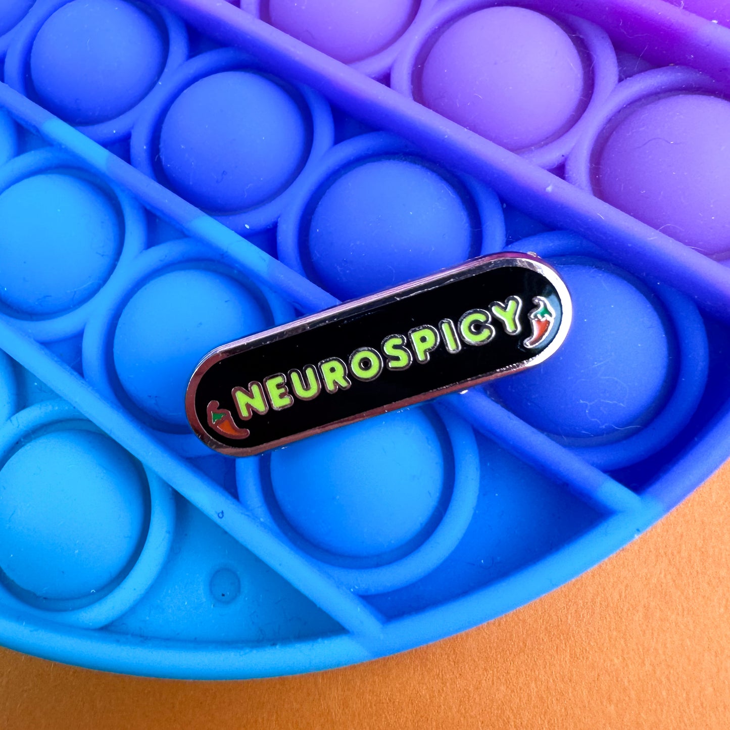 A capsule shaped enamel pin that reads "Neurospicy" with chili peppers around it on a pop it fidget toy.
