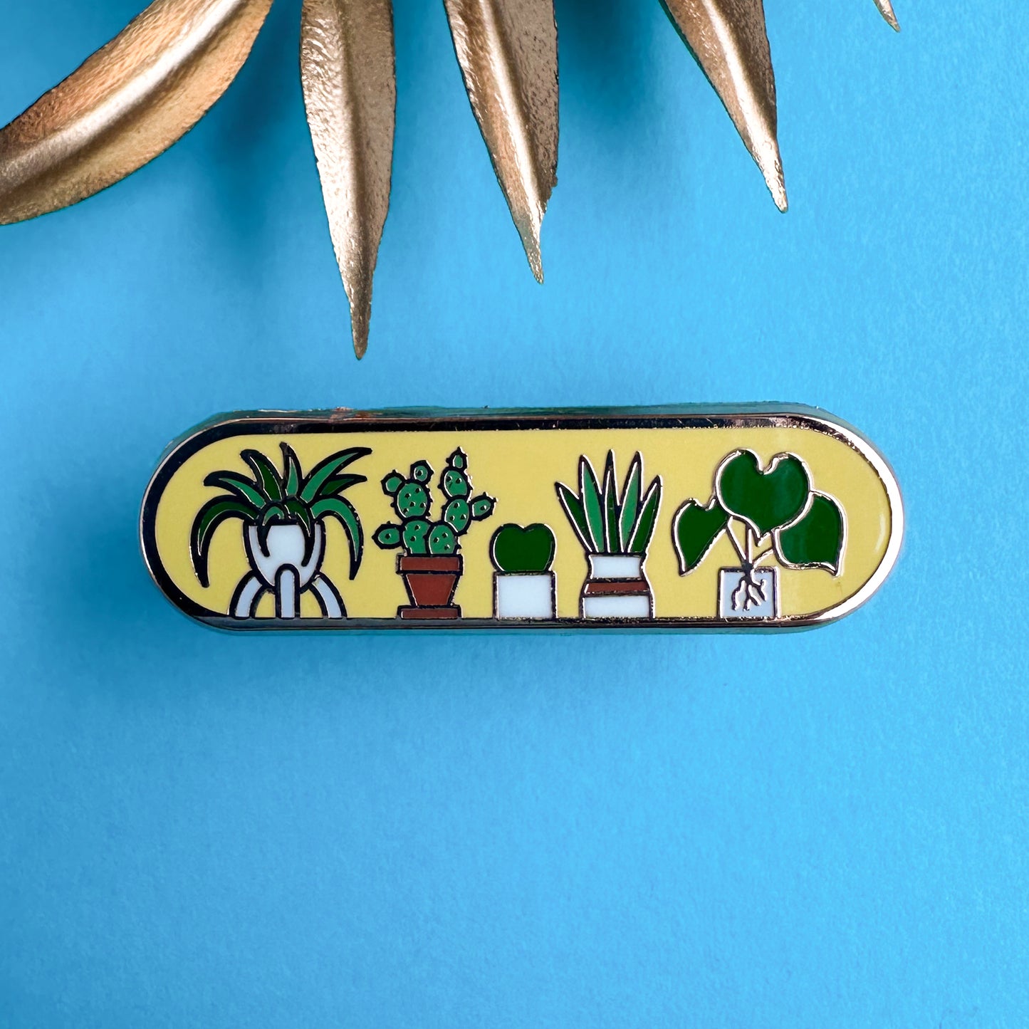 An oval shaped pin with illustrations of cute house plants like a cactus, spider plant, Hoya, and snake plant on it. The pin is on a blue background with some gold leaves in the background.