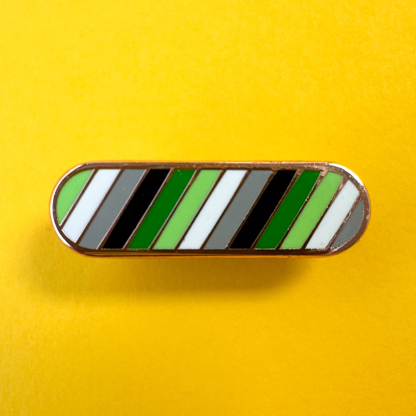 A bandaid shaped pin with diagonal bands of color in the colors of the Aromantic Pride Flag, green, light green, white, grey and black. The pin is on a yellow background.