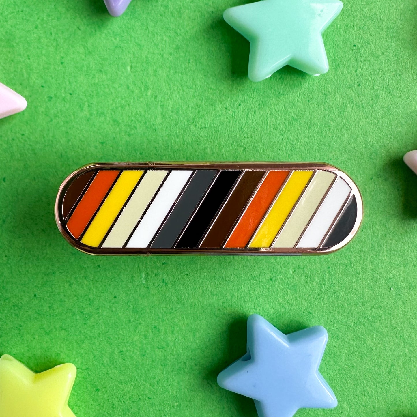 A capsule shaped pin in the colors of the Bear Pride flag in diagonal stripes. The pin is on a green paper background with plastic star beads around it.