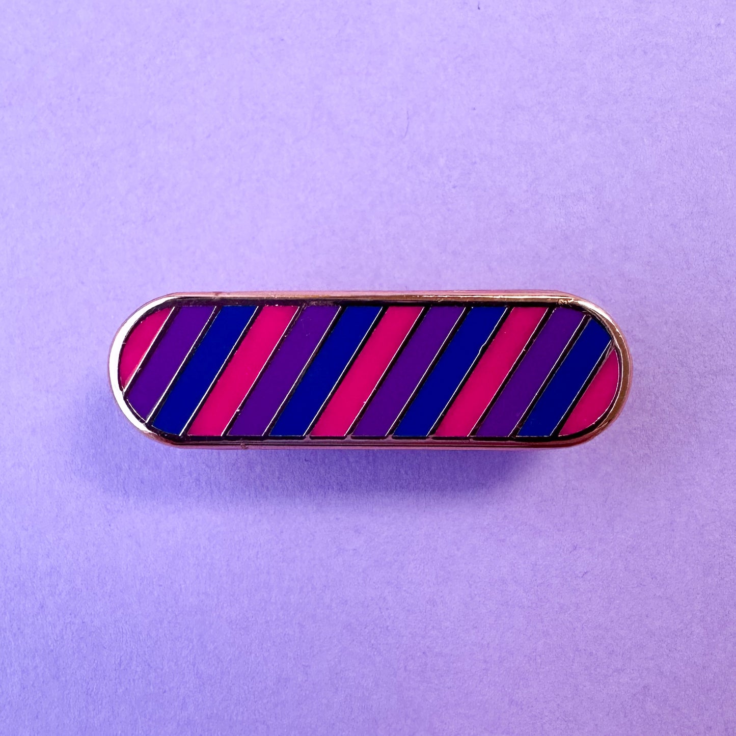 A bandaid shaped enamel pin with diagonal stripes in pink, purple, and blue which are the colors of the Bi Pride Flag. The pin is on a lavender background.
