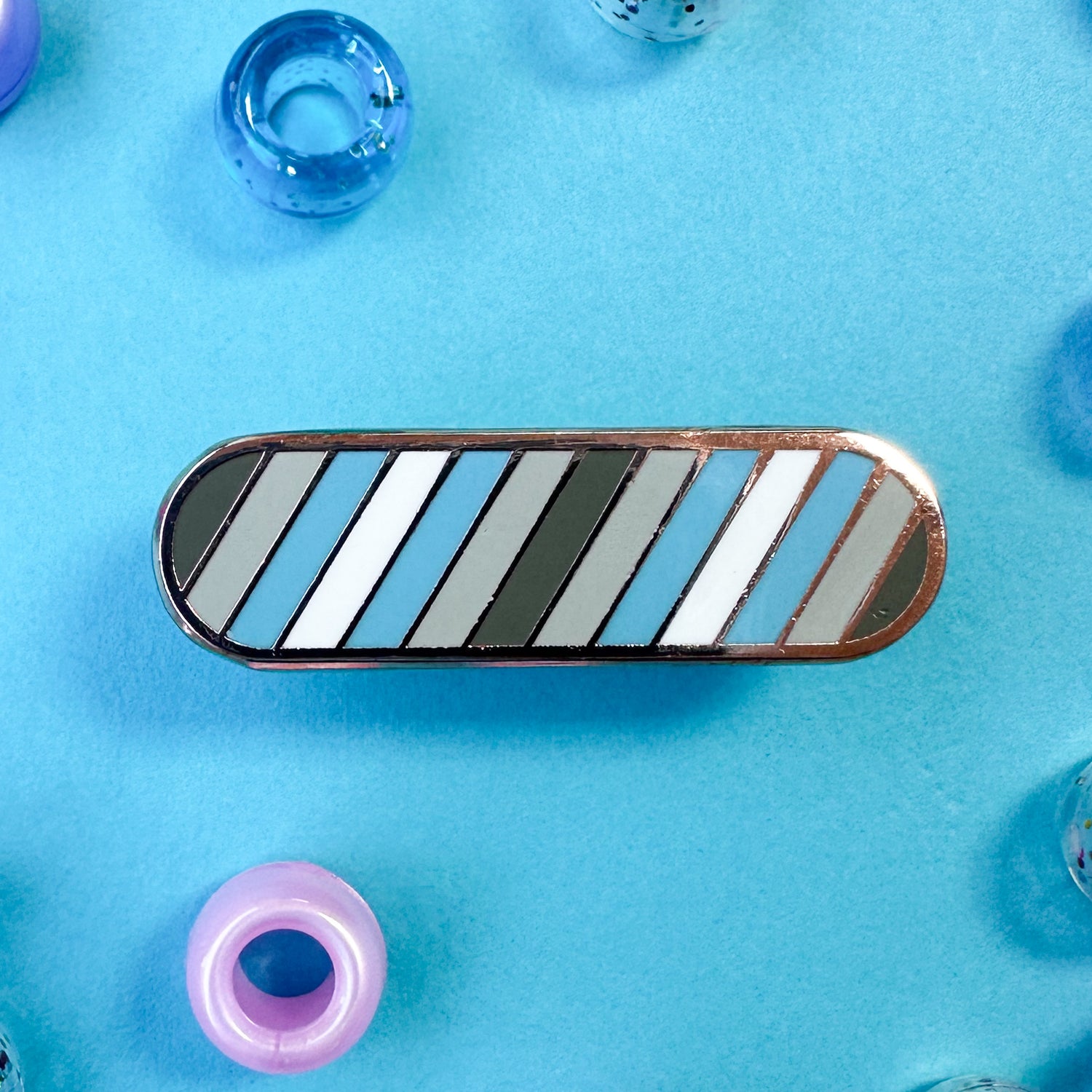 A bandaid shaped pin with diagonal stripes in grey, light grey, light blue, and white, which are the colors of the Demiboy Pride flag.  The pin is on a blue background with pony beads around it.