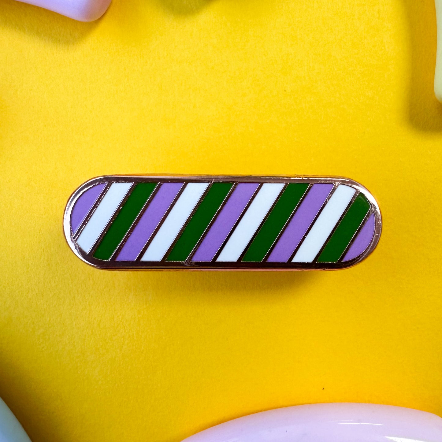 A capsule shaped enamel pin in the colors of the genderqueer pride flag on a yellow background with beads around.