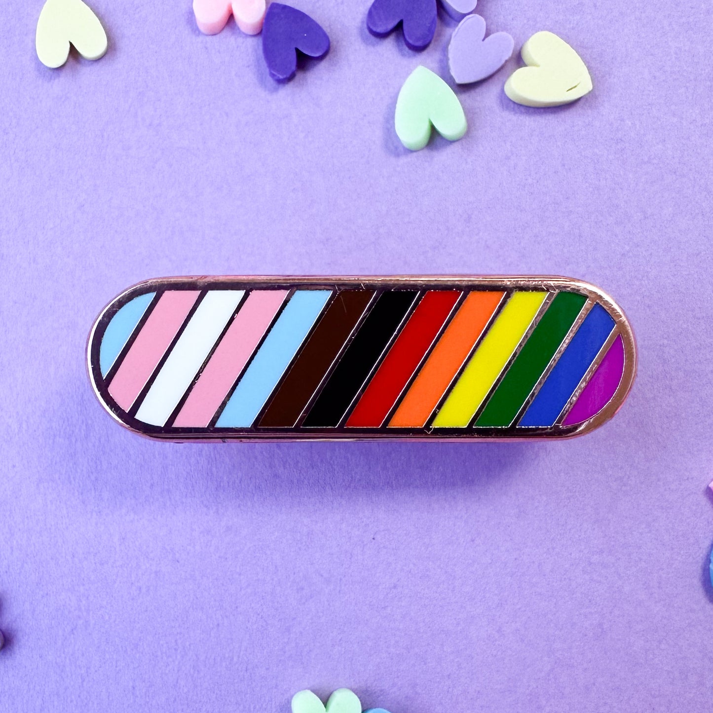An oval shaped pin with diagonal stripes in the colors of the progress pride flag. The pin is on a lavender background with pastel heart confetti around.