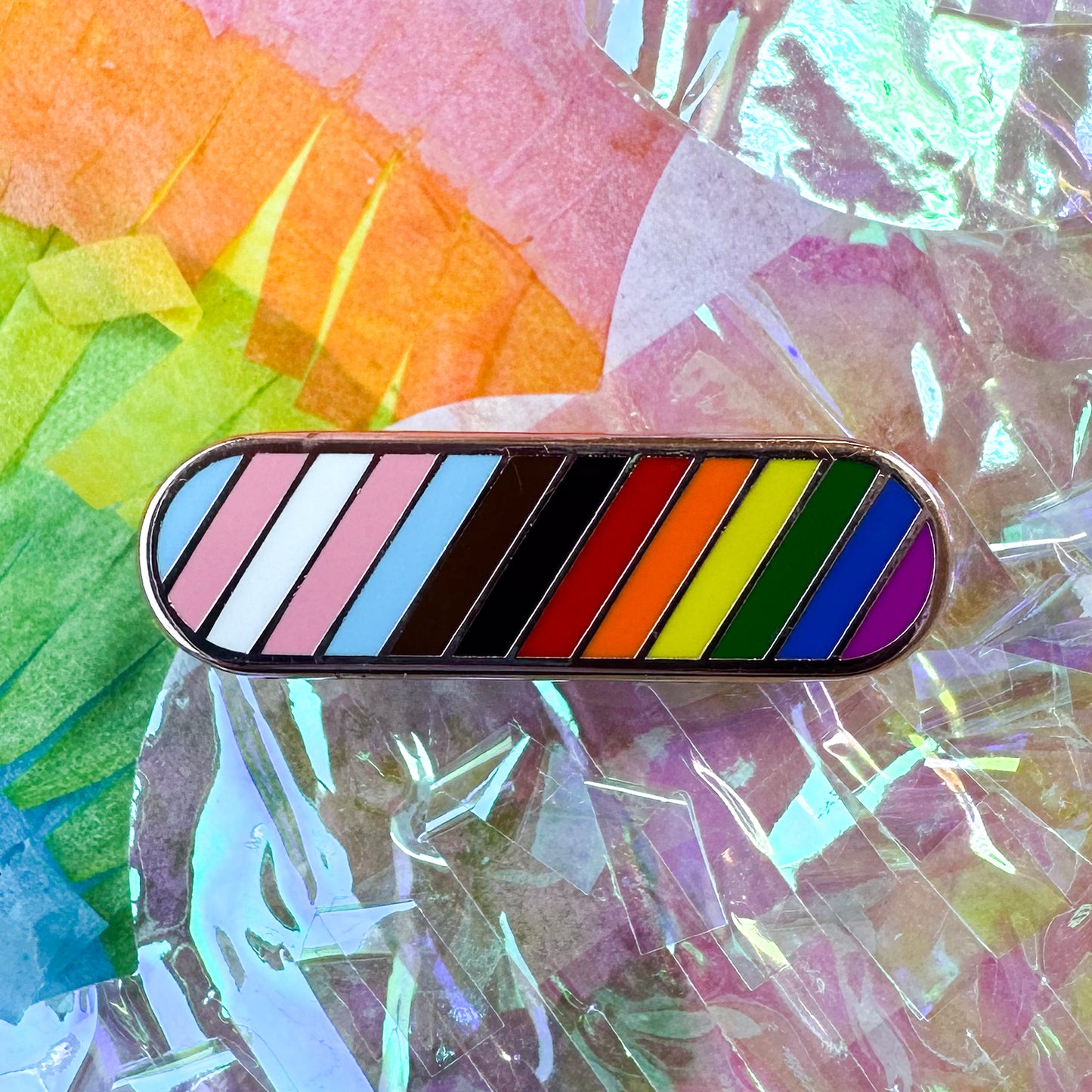 A capsule shaped pin with diagonal stripes in the colors of the inclusive pride flag. The pin is on top of a pastel rainbow piñata. 