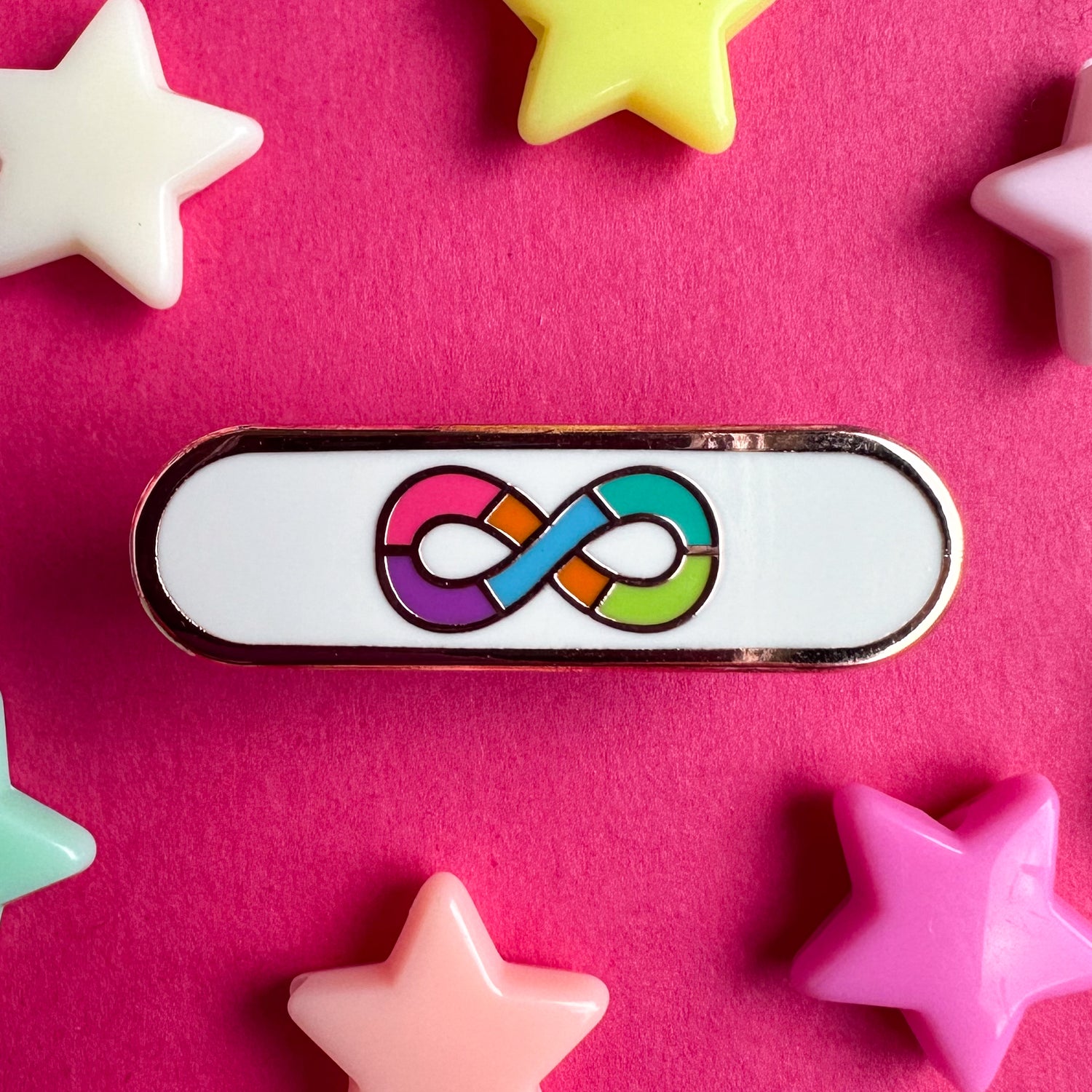 An enamel pin with the rainbow infinity symbol of the Neurodivergent Pride Flag on a white background in a capsule shape. The pin is on a pink background with pastel star beads around it.