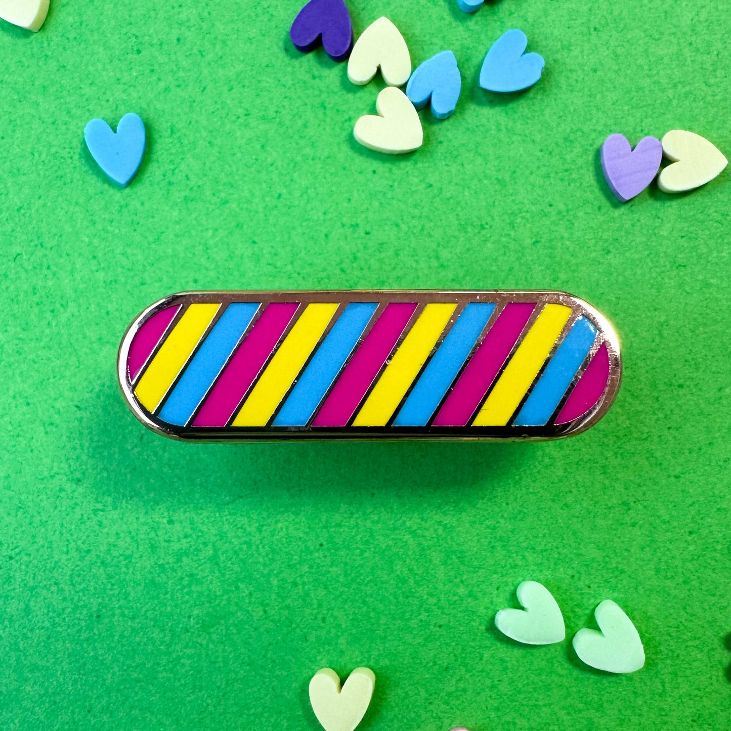 An oval shaped pin in the Pan Pride flag colors. The pin is on a green background with heart confetti around.