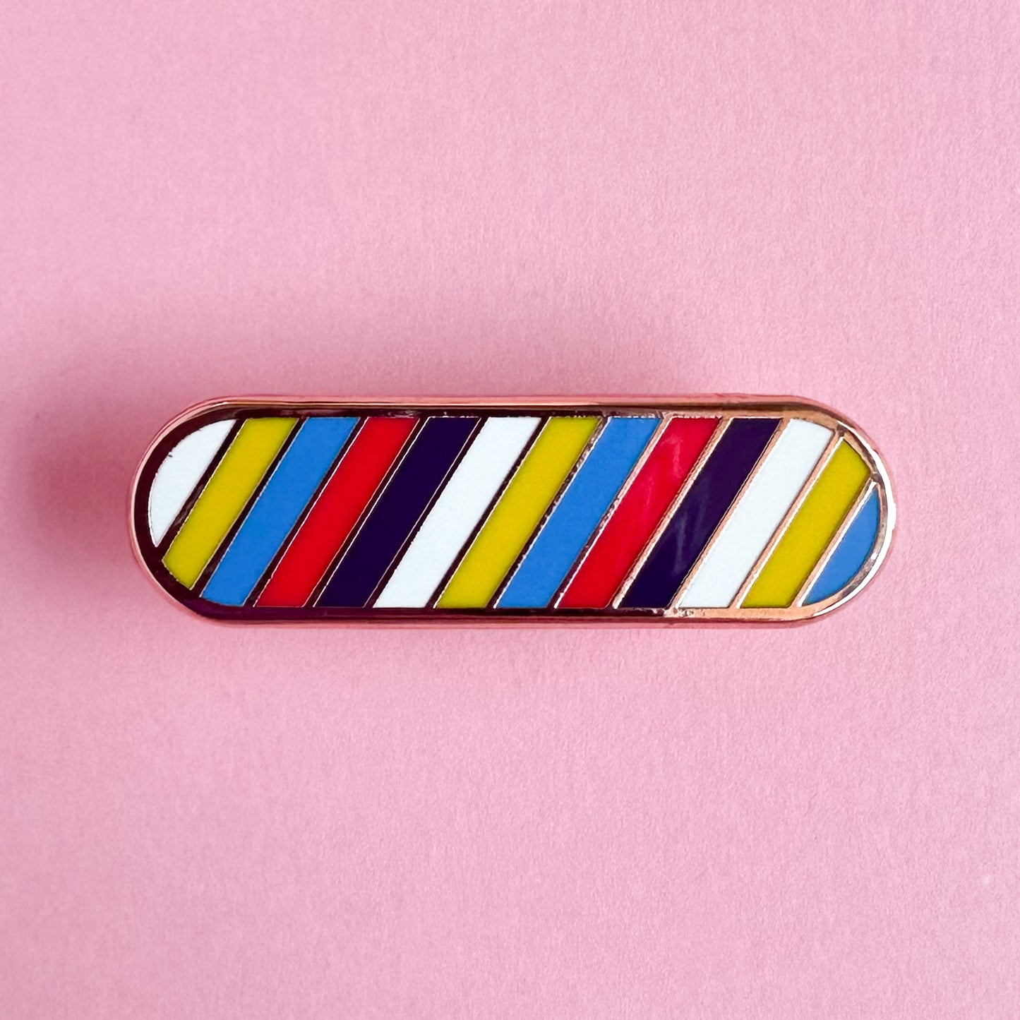 An oval shaped enamel pin in the colors of the new Polyamorous Pride Flag in diagonal stripes across it. The pin is on a pink paper background.