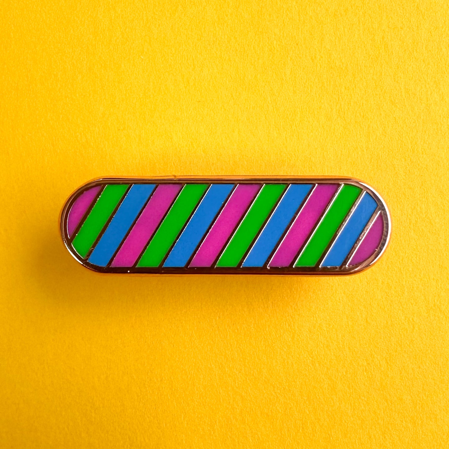 A capsule shaped enamel pin in the colors of the Polysexual pride flag which are pink, green, and blue. The pin is on a yellow paper background.