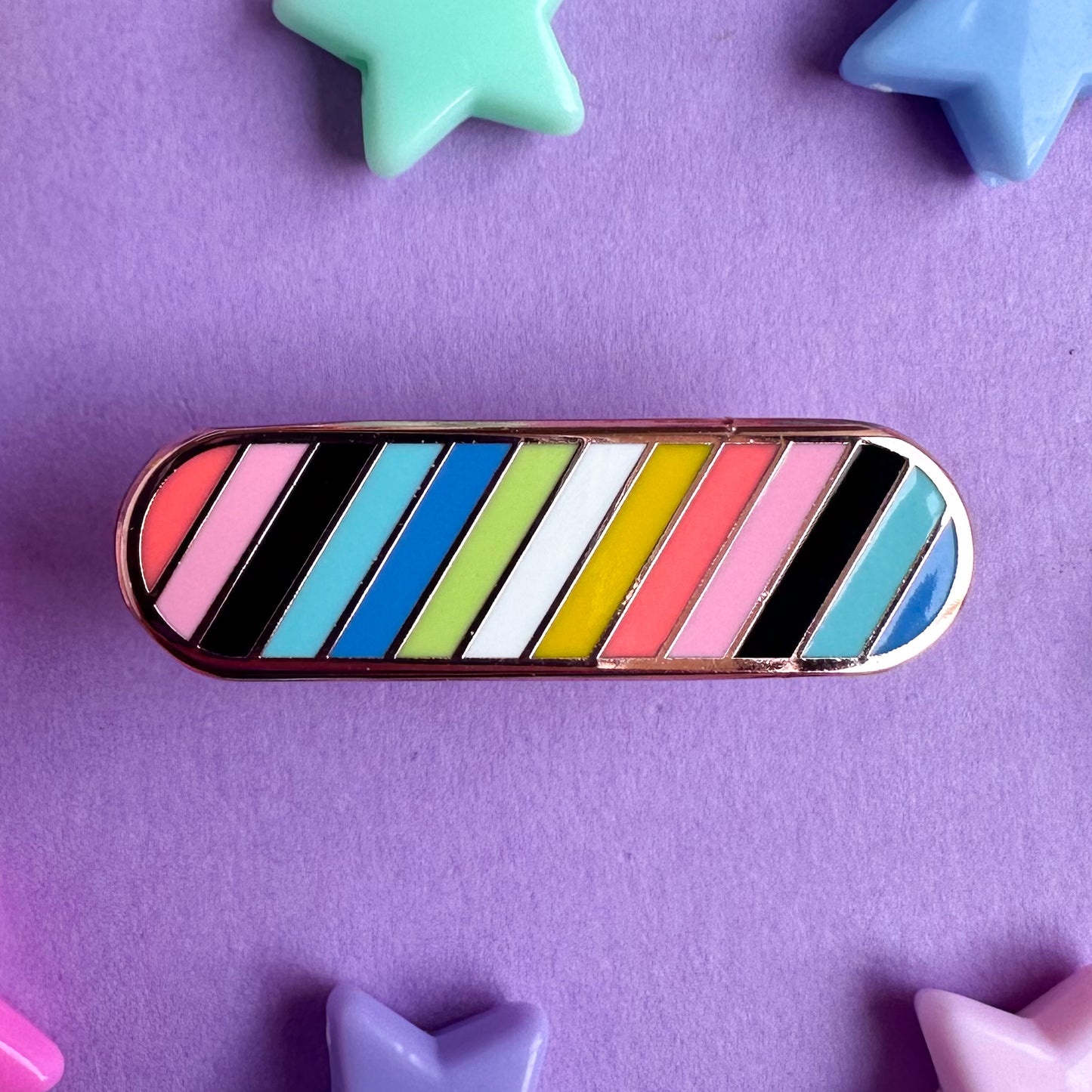 An enamel pin shaped like a bandaid in the colors of the Queer Pride Flag with diagonal stripes. The pin is on a purple background with plastic star beads around it.