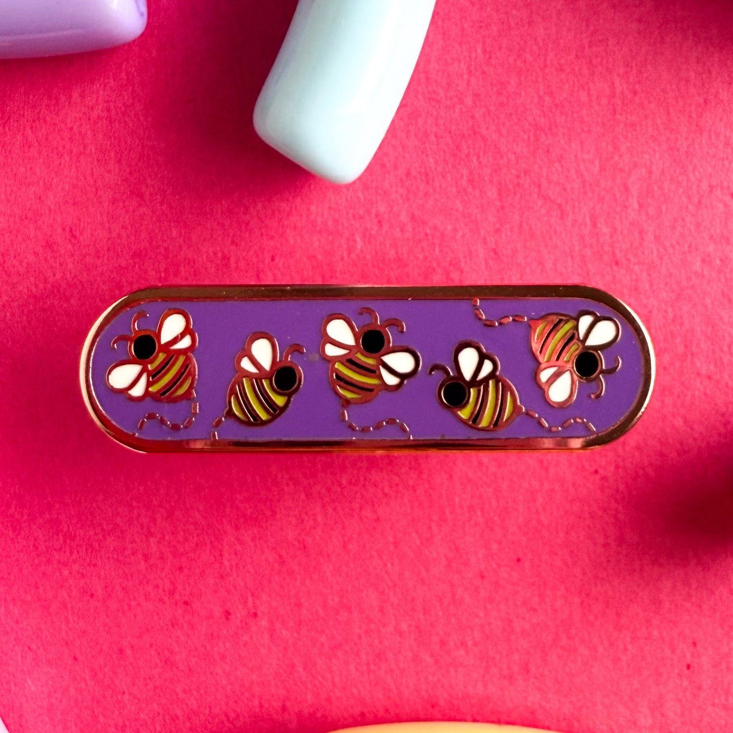 A bandaid shaped enamel pin with five little bees flying around. The pin is on a pink background.