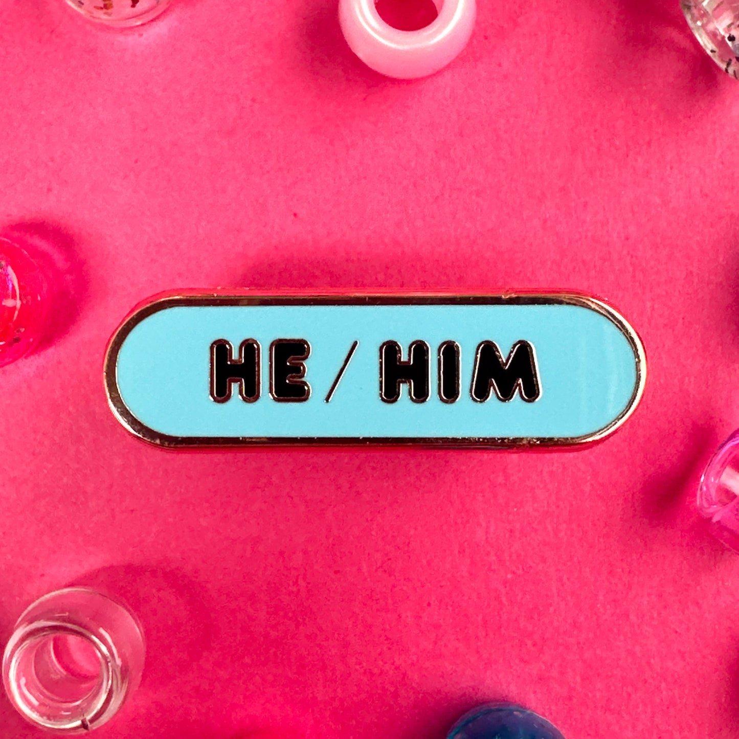 A blue oval shaped pin with the words "He/Him" in rounded letters. The pin is on a hot background with pony beads around it.