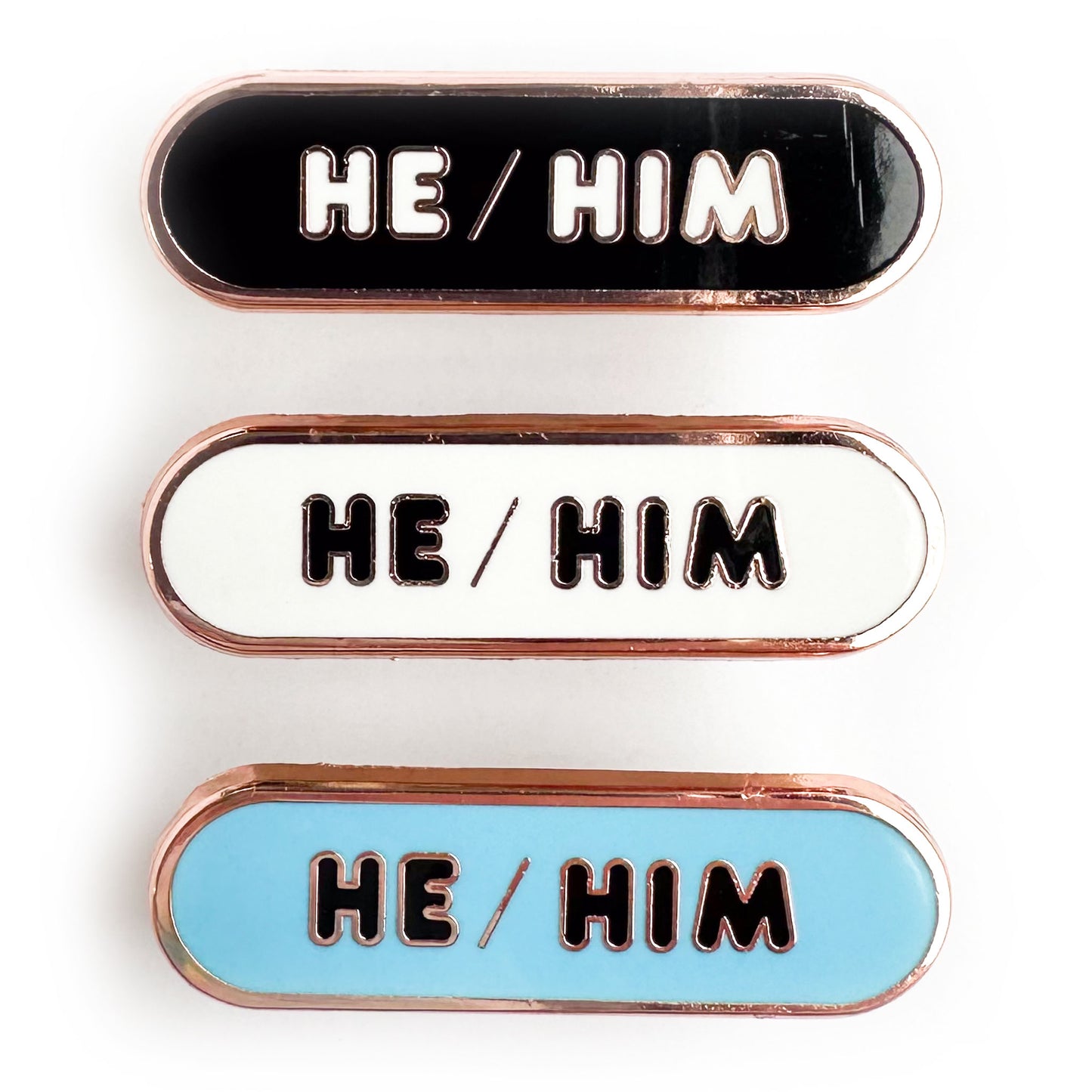 Three capsule shaped pins that all have the words "He/Him" in bubble letters. One of the pins is black, one is white and one is light blue.