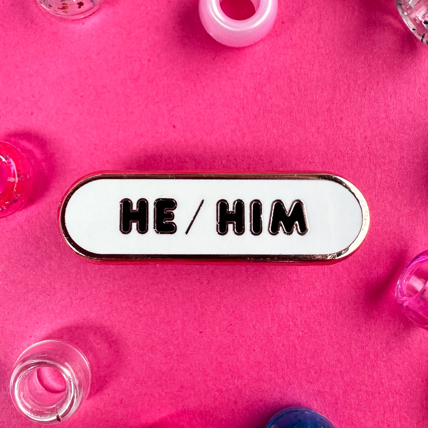 A white capsule pin with bubble letters that read "He/Him". The pin is on a hot pink background with pony beads around it.