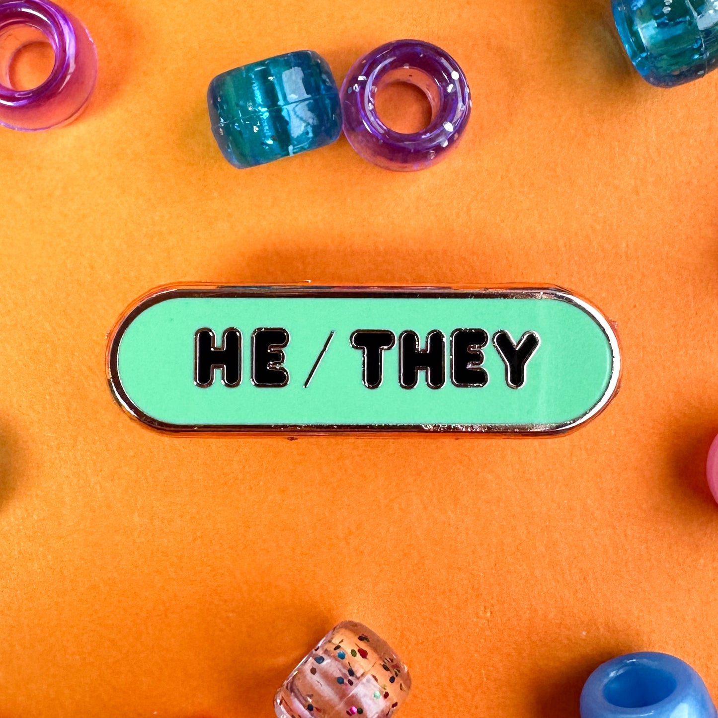 A mint oval shaped pin with black puffy letters that read "He/They" the pin is on an orange background with pony beads around.